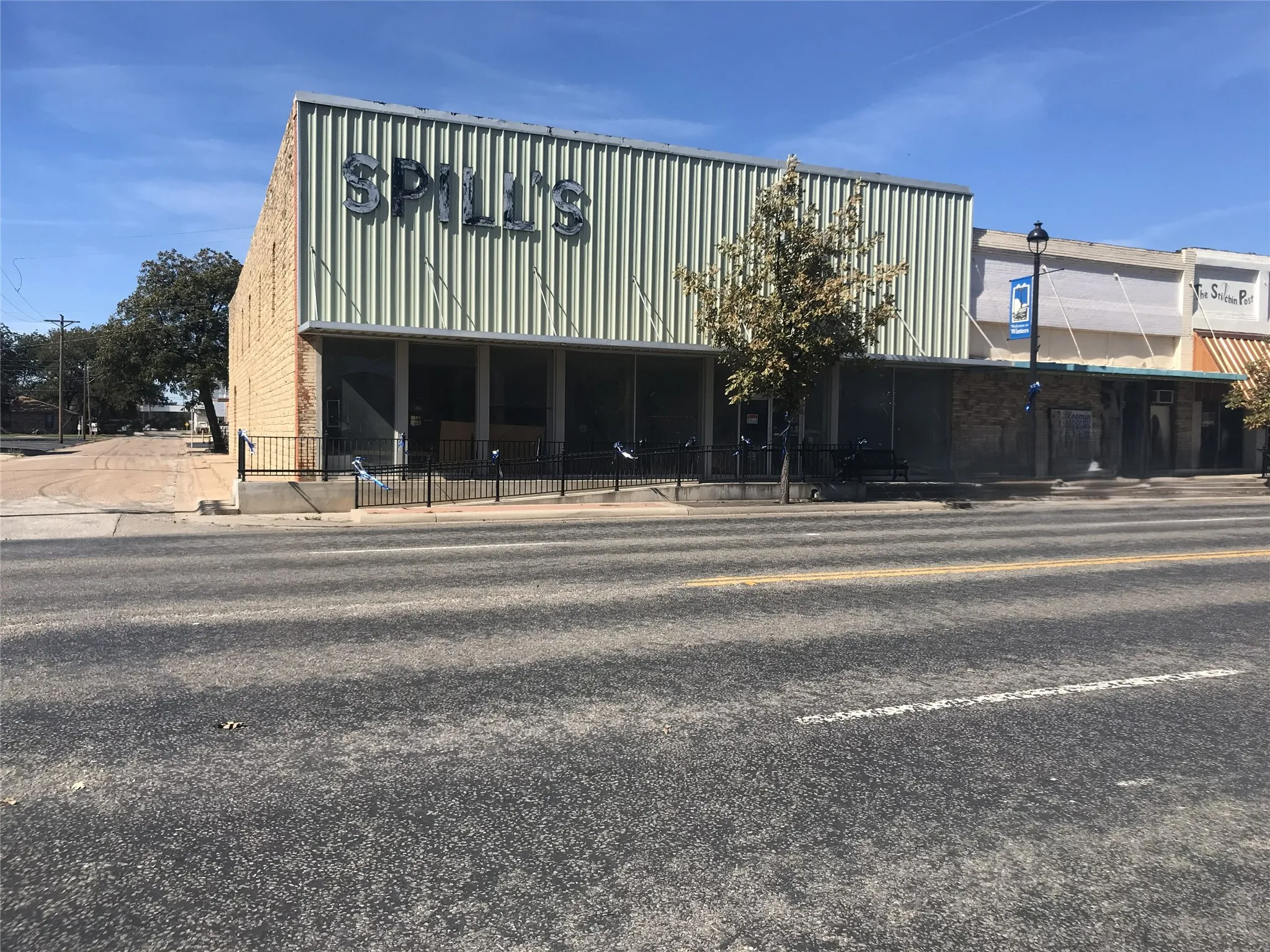 Brick facade with a corrugated metal upper section, large storefront windows, and a black wrought iron fence