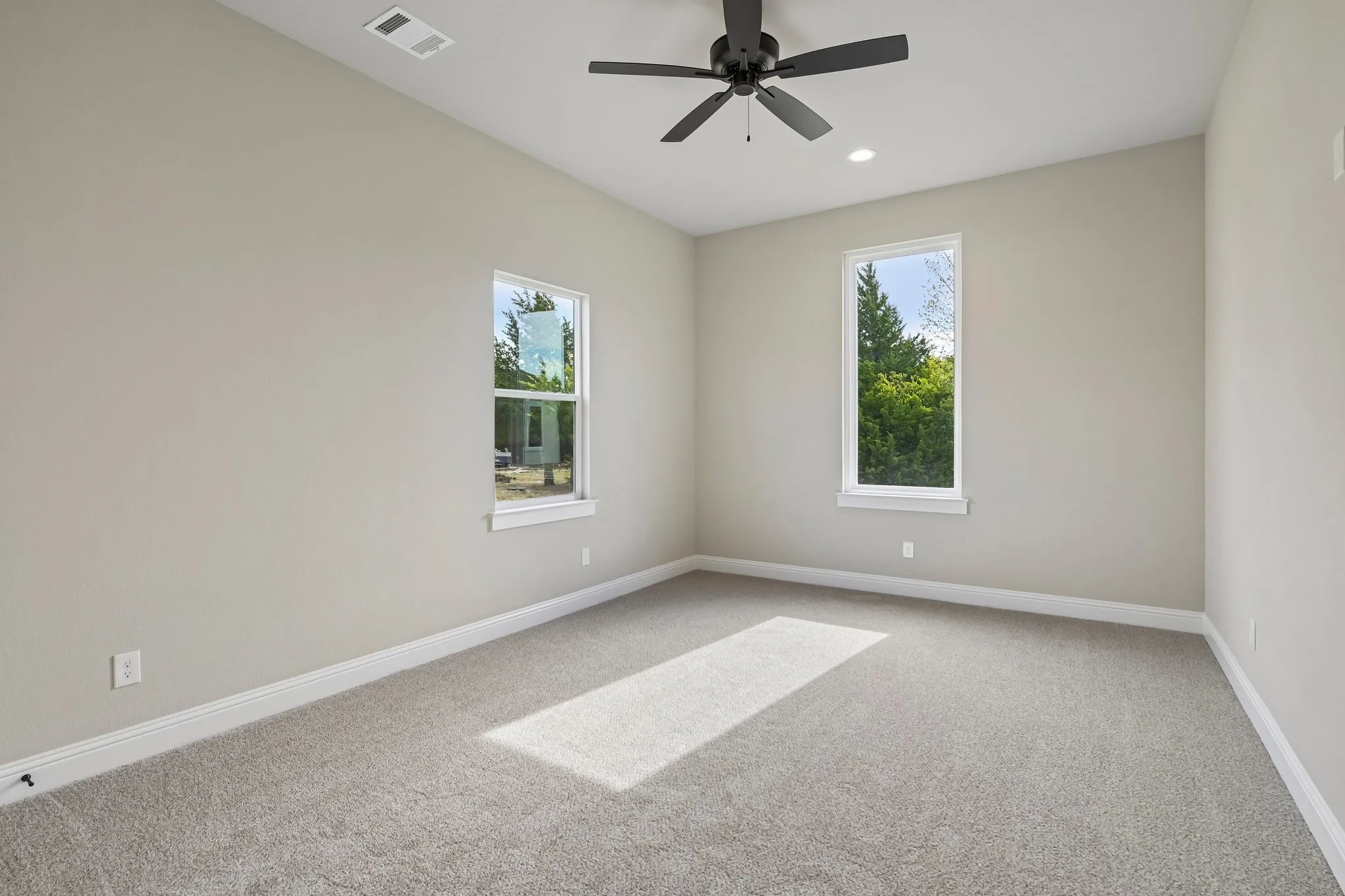 Bright, airy bedroom showcasing dual windows with natural light, neutral wall finishes, neutral carpet flooring, and a ceiling fan with recessed lighting