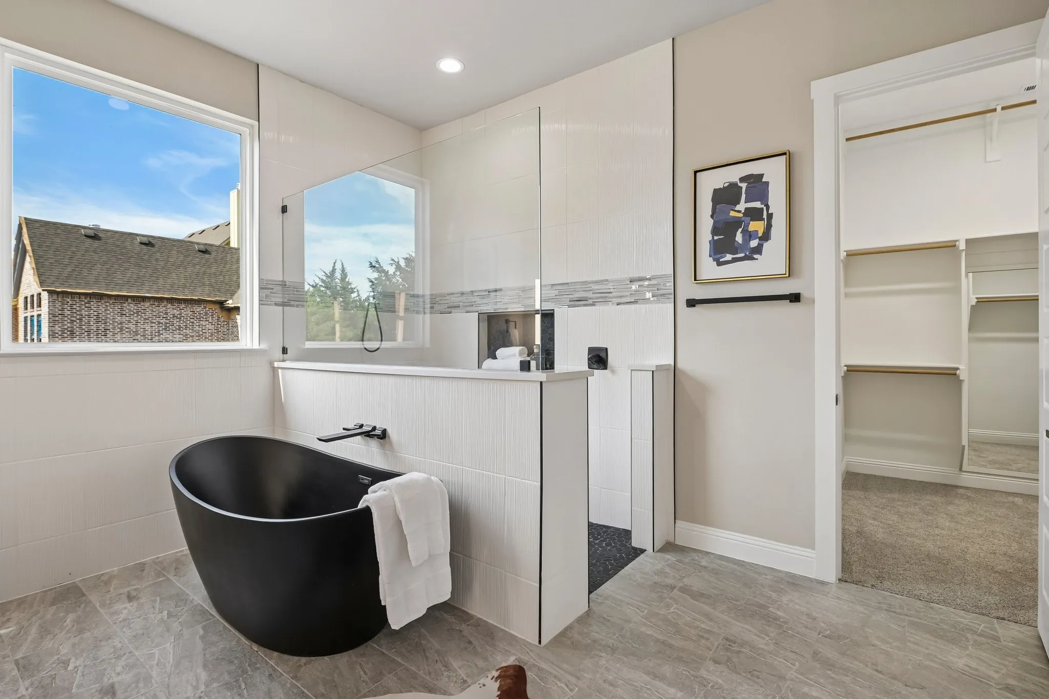 Freestanding matte black bathtub with a wall-mounted faucet, separate shower enclosure with glass panels, light-toned tile walls, and stone-look flooring