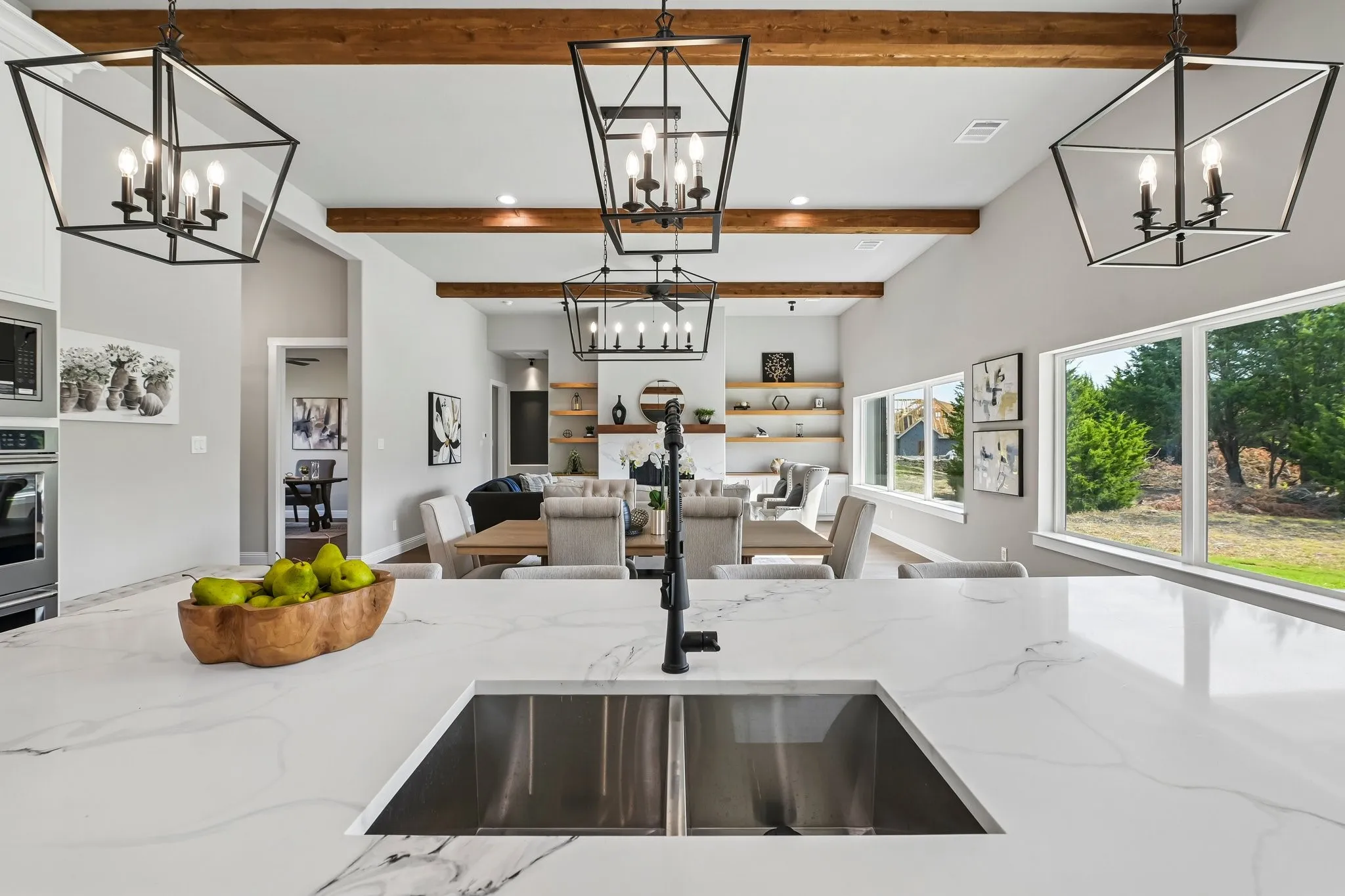 Open-concept kitchen and living area featuring exposed wood beams, vaulted ceilings, a large island with an undermount double-basin sink, and expansive​​‌​​​​‌​​‌‌​‌‌​​​‌‌​‌​‌​‌​​​‌​​ windows