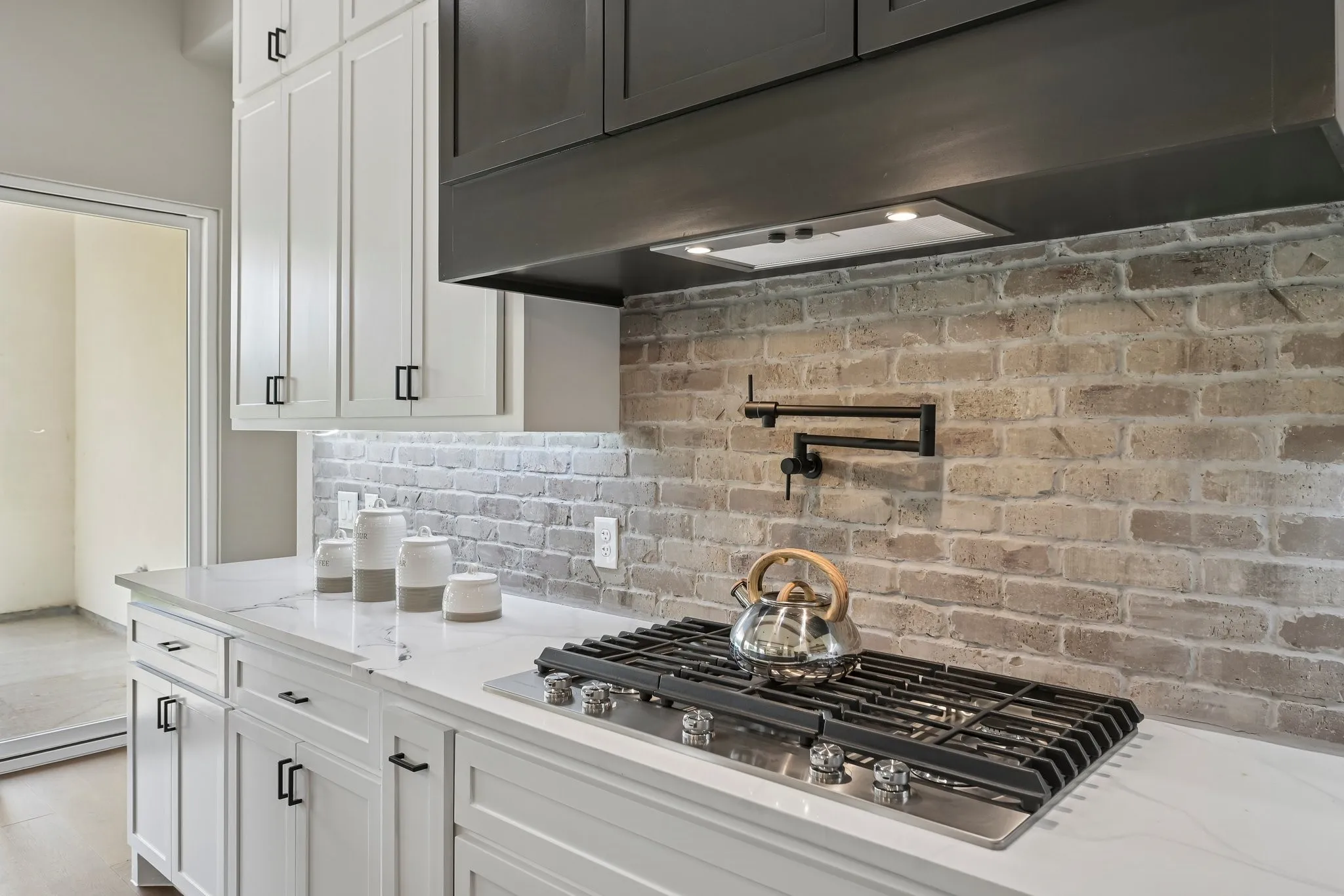 White shaker-style cabinetry with matte black hardware, stone-look countertops, a stainless steel gas cooktop, a brick-style backsplash, and a black pot filler