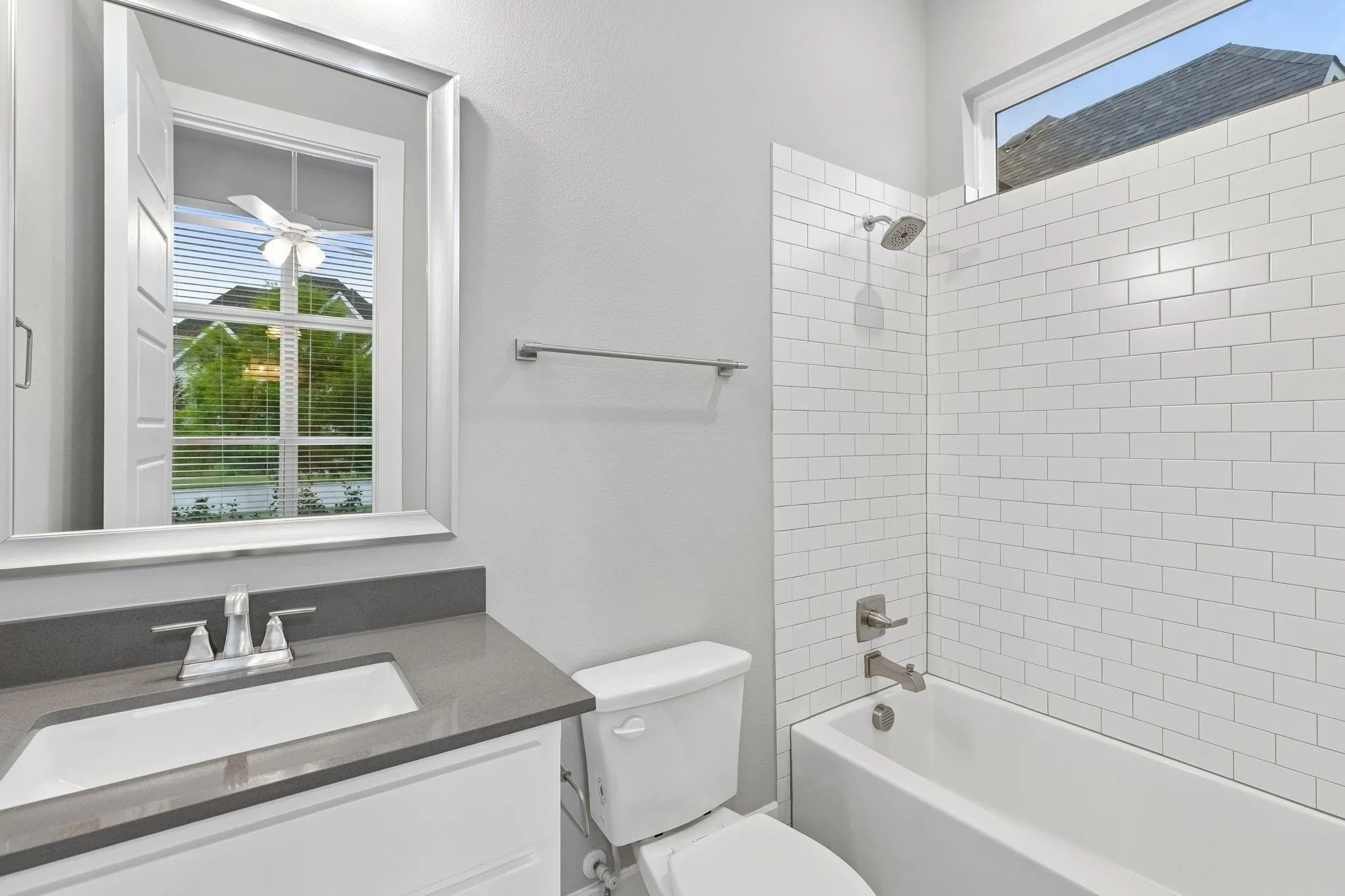 White vanity with a gray countertop and undermount sink, a framed mirror, a toilet, a bathtub with a shower, and subway tile surround