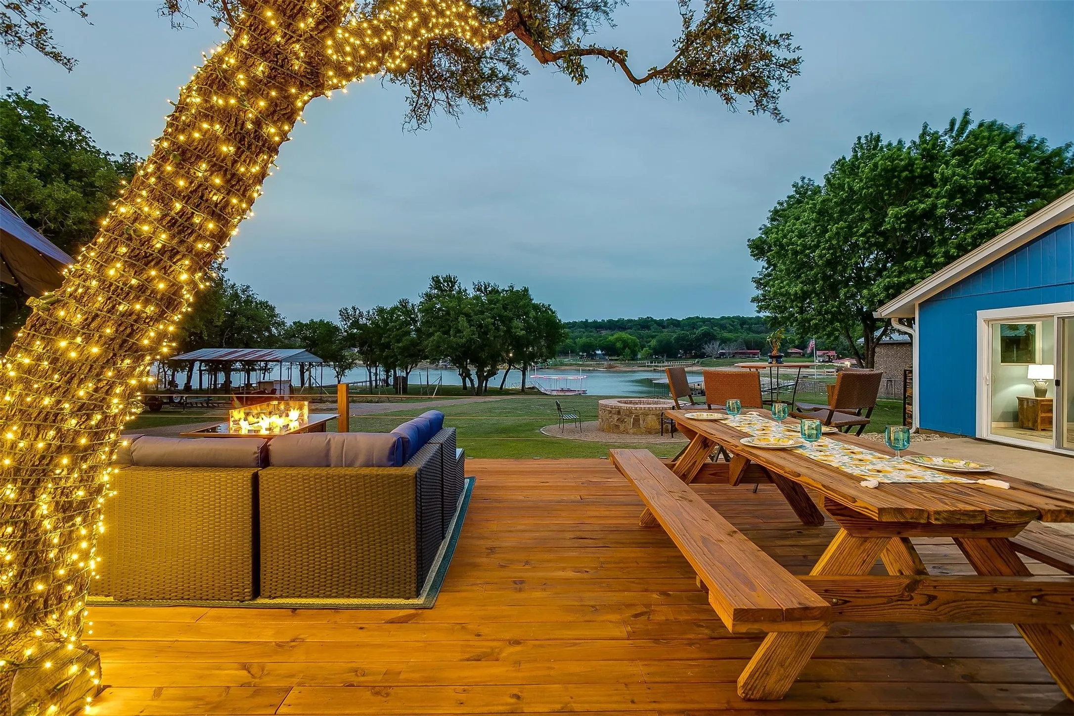 Wood-finish deck with a built-in fire pit and a large picnic table, overlooking a body of water