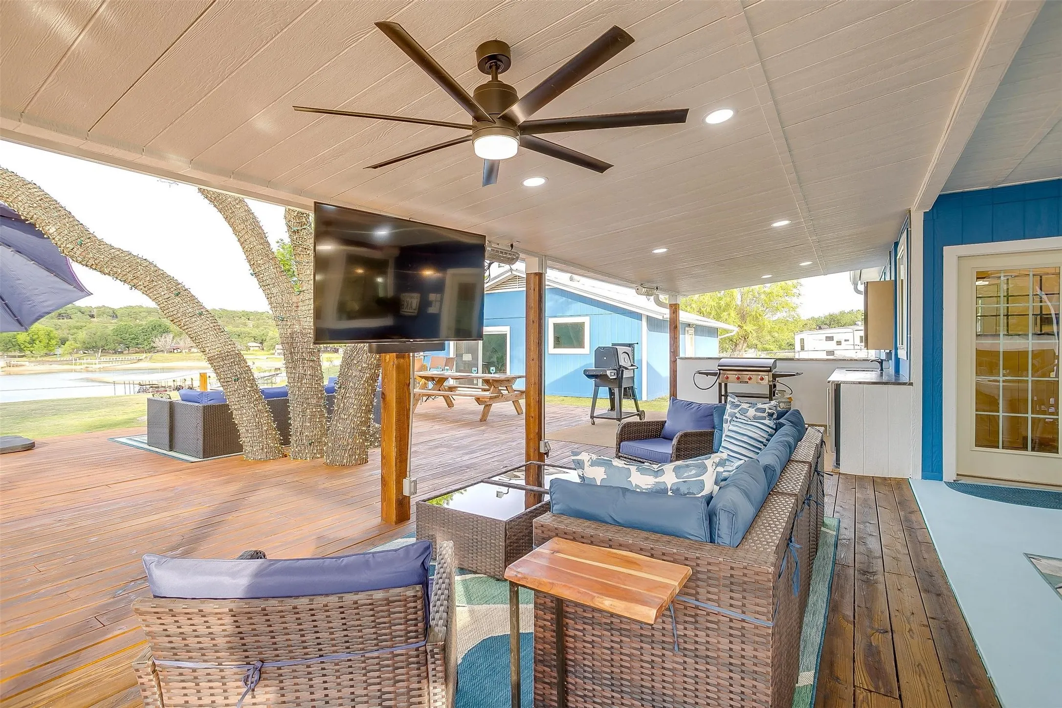 Expansive outdoor deck with wood-finish flooring, featuring a large ceiling fan with integrated lighting, recessed lighting, and a built-in outdoor kitchen area