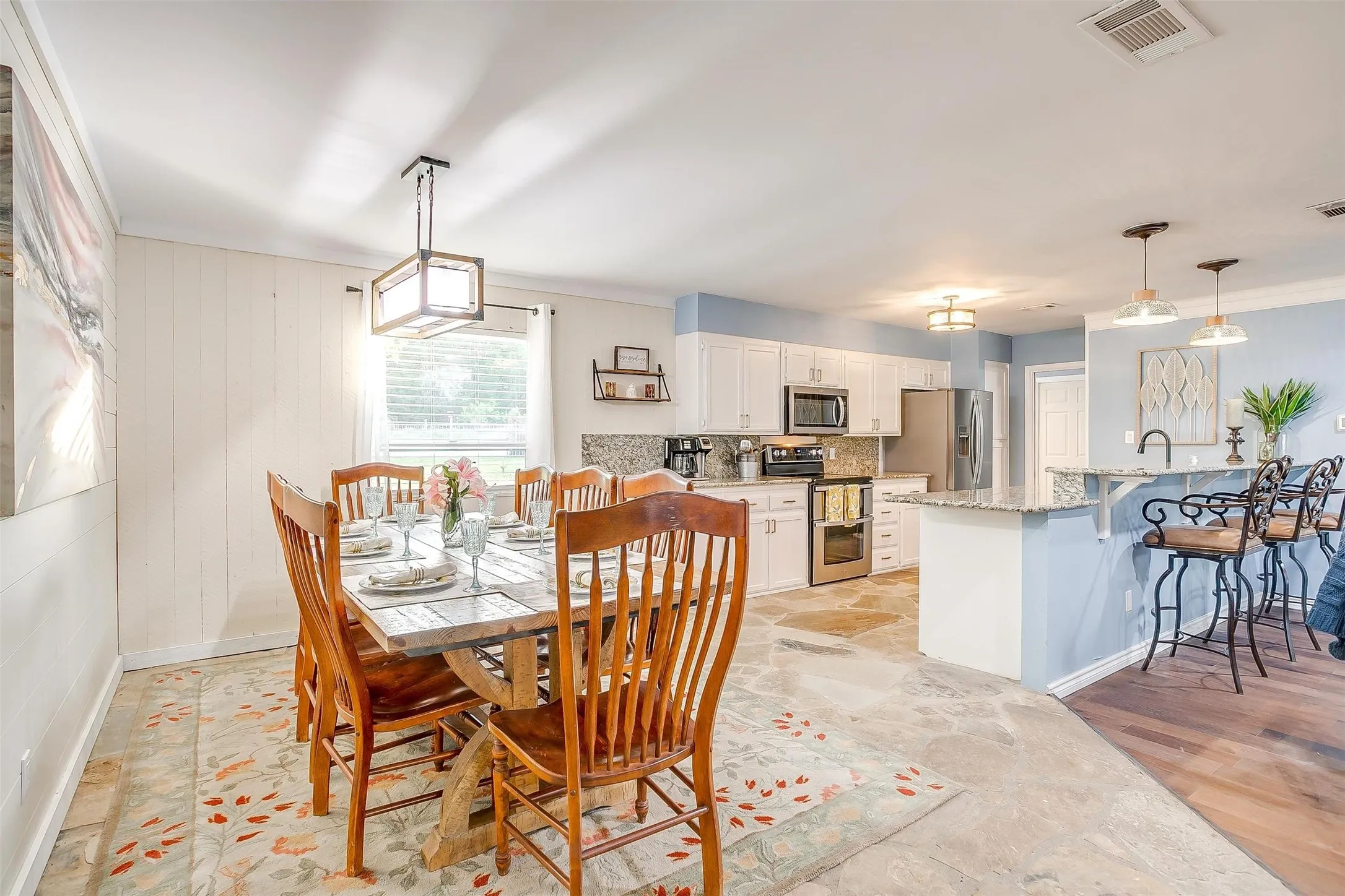 Open-concept dining and kitchen space featuring stone flooring, a contemporary dining chandelier, and a kitchen with white cabinetry, stainless steel appliances, and a breakfast bar with pendant lighting