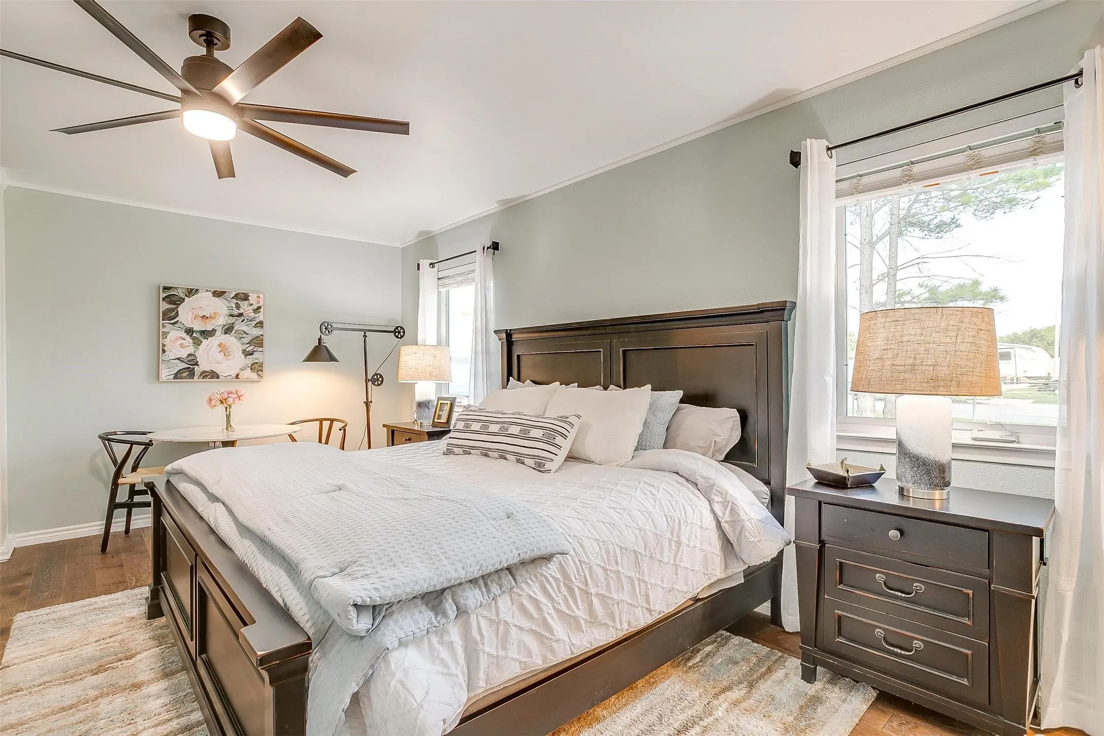 Wood-finish flooring, ceiling fan with integrated lighting, multiple windows with roller blinds and curtains, and crown molding
