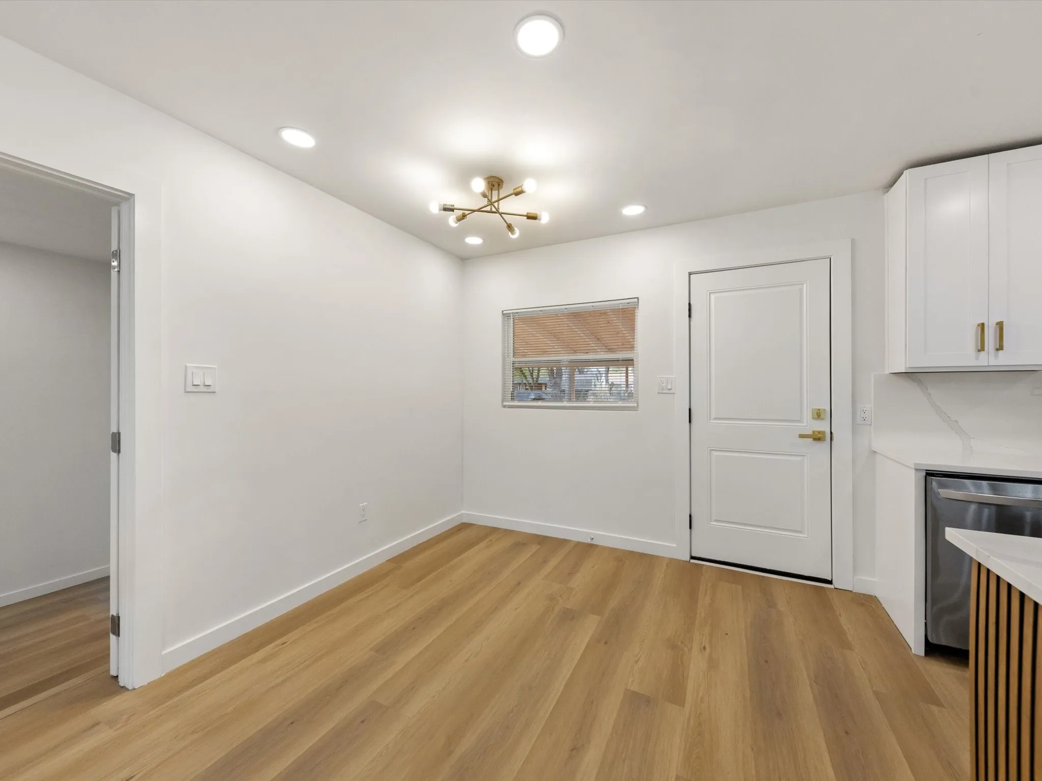 Recessed ceiling lighting, wood-finish flooring, contemporary ceiling fixture, white paneled door, and a kitchen area with white cabinetry