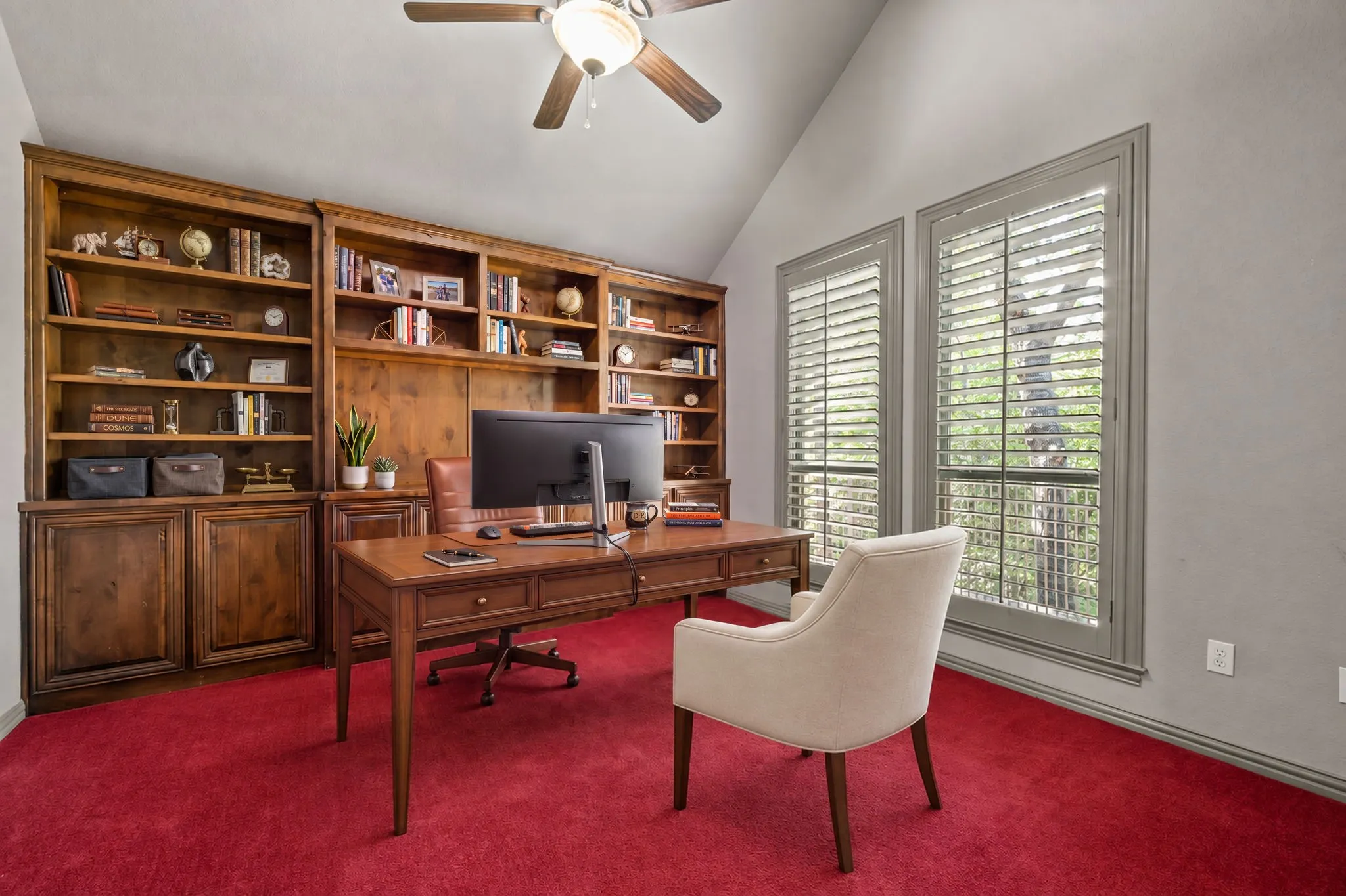 Large office with  built-in custom wood cabinetry, multiple windows with plantation shutters, ceiling fan with integrated lighting, and red carpet flooring