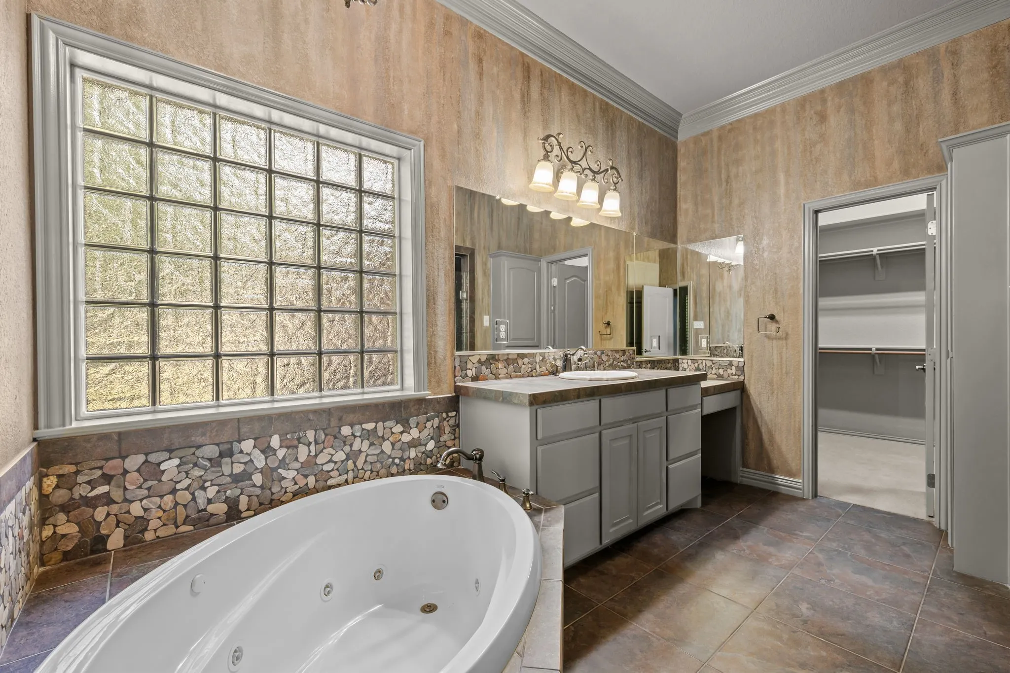 Jetted soaking tub, glass block window, dual vanity with integrated seating, stone flooring, and a walk-in closet