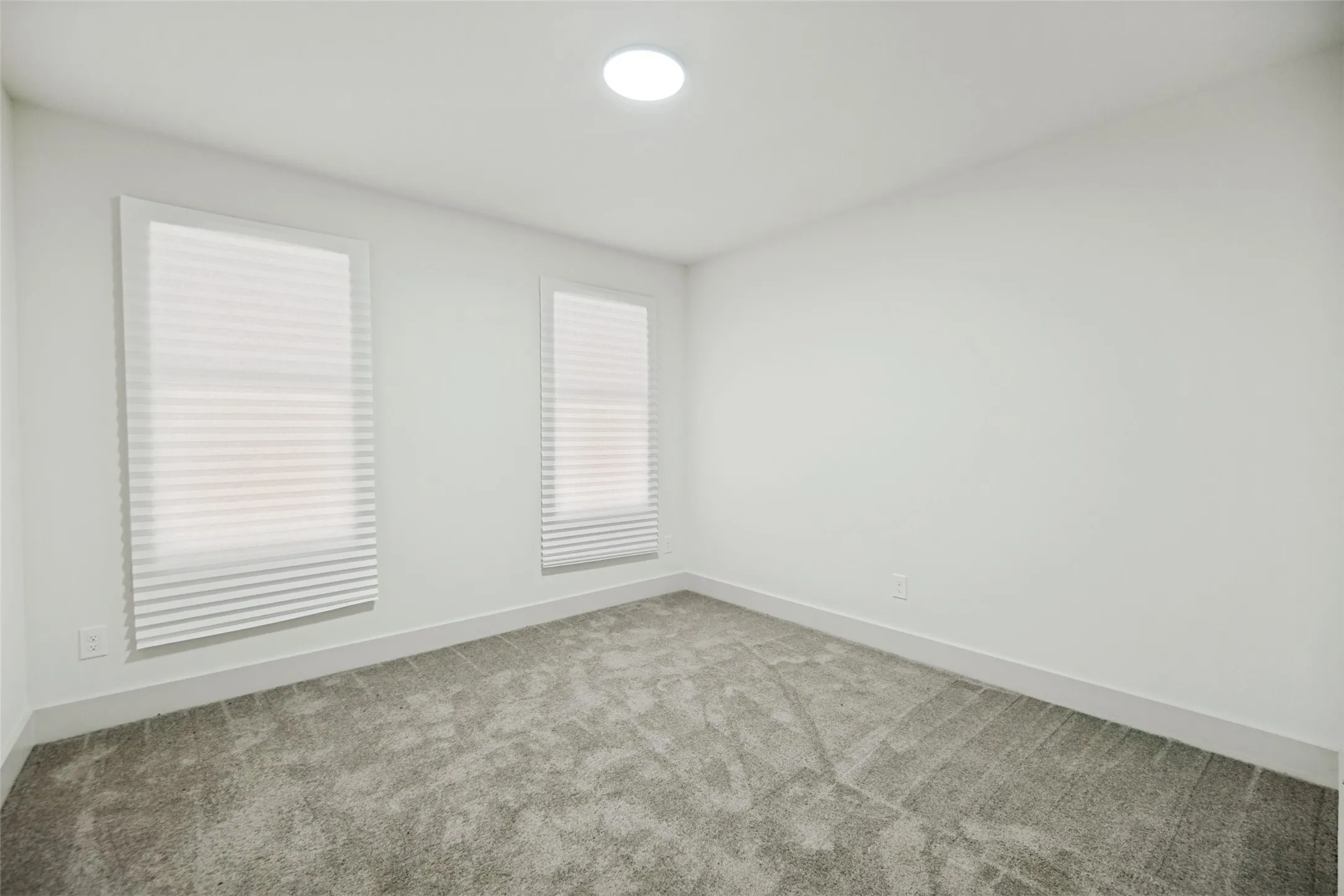 Bright interior featuring white walls, gray textured carpet, two rectangular windows with white blinds, and a recessed ceiling light fixture