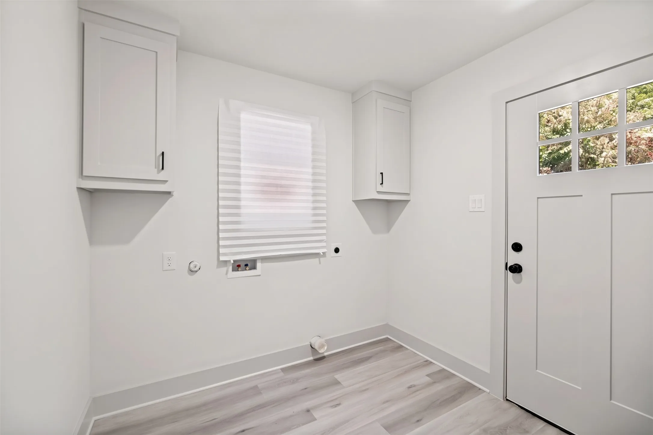 Light-toned wood-finish flooring, white paneled door with glass inserts, two wall-mounted white cabinets, and a window with horizontal blinds