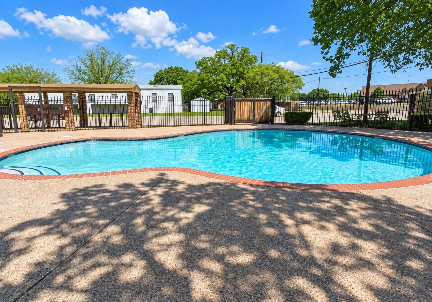 Curved swimming pool with a textured concrete deck, brick coping, and a surrounding black metal fence