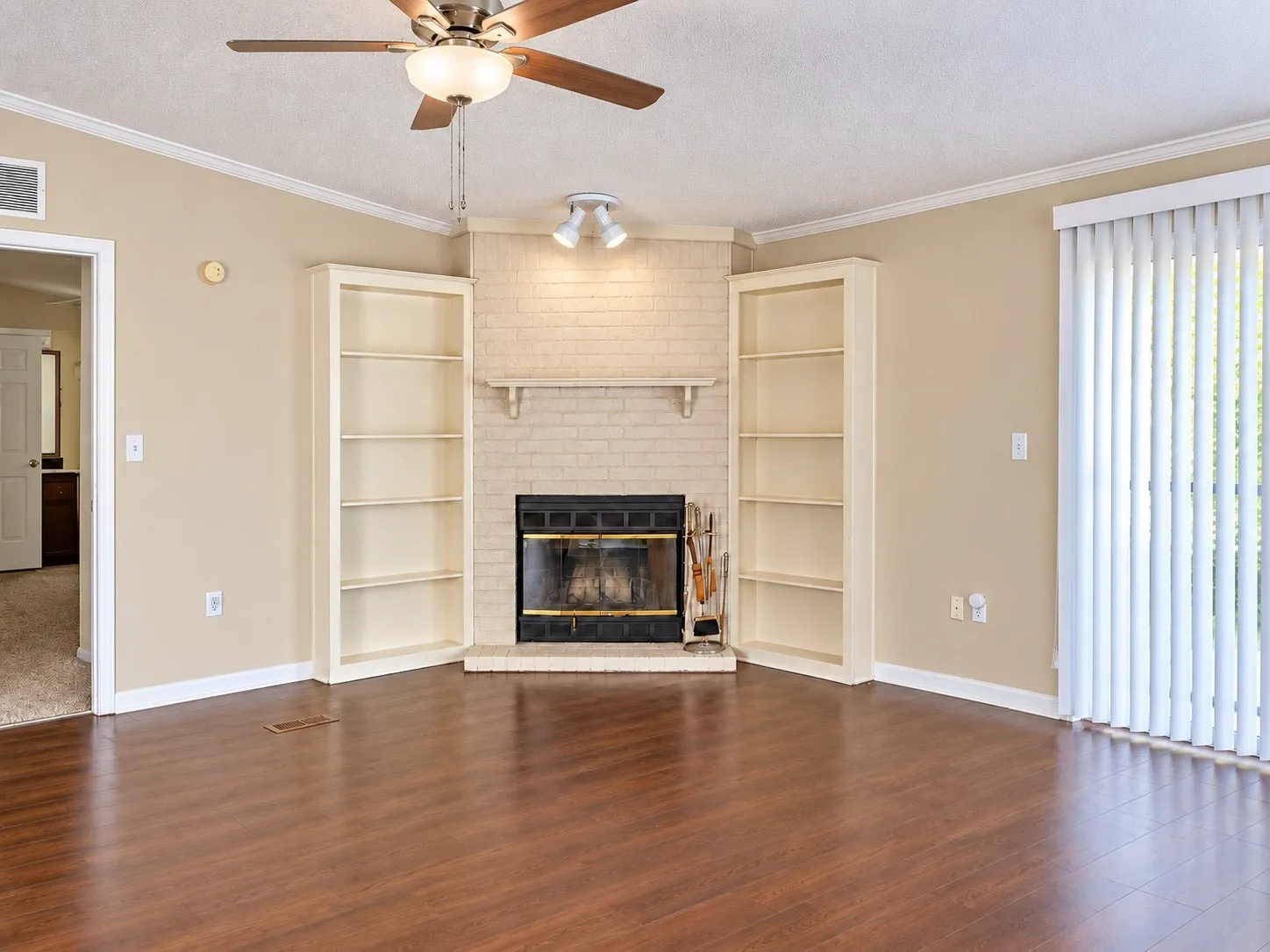 Corner fireplace with painted brick surround, built-in shelving, wood-finish flooring, ceiling fan, and sliding glass door
