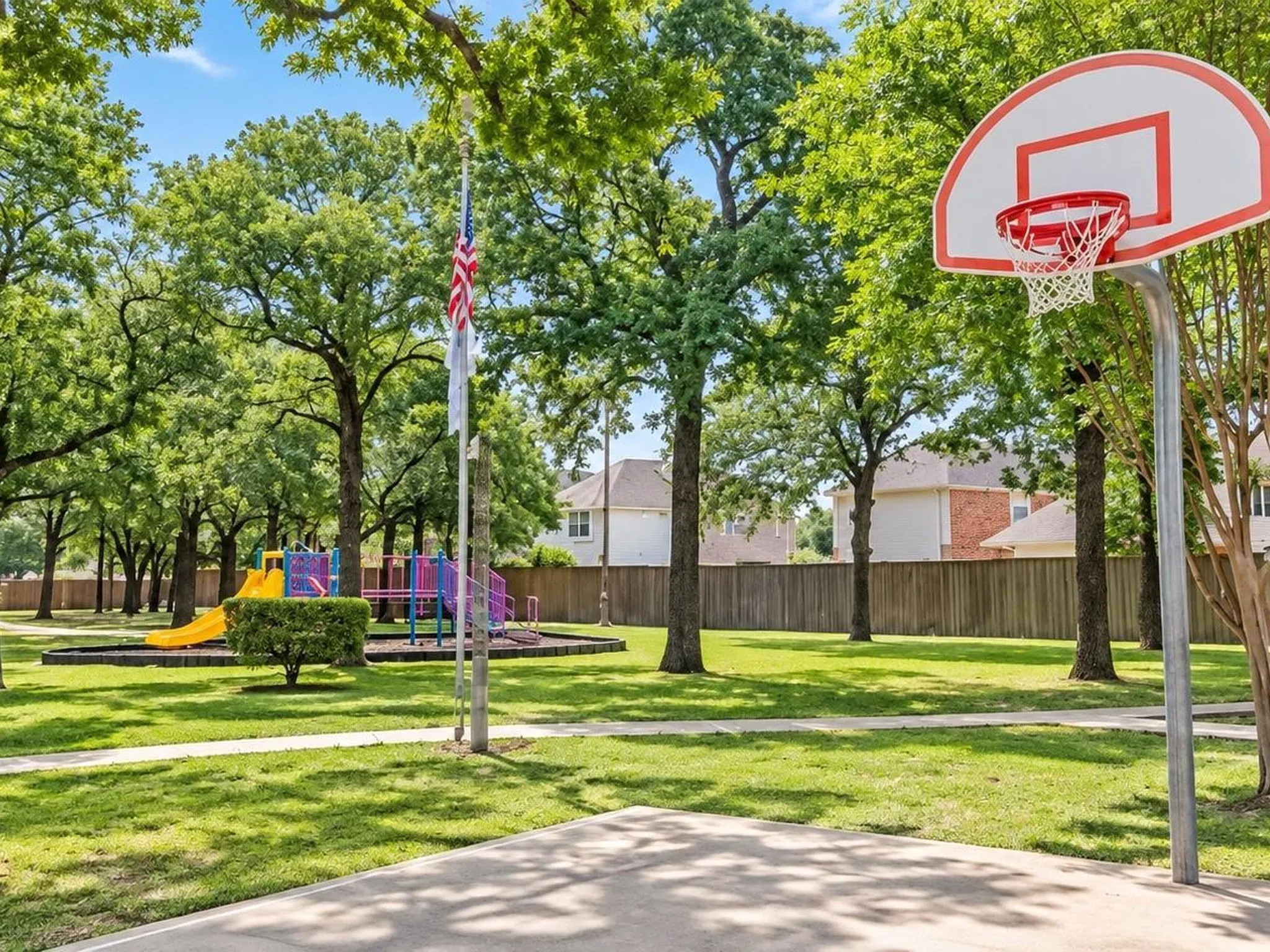 Outdoor recreational space featuring a basketball court, playground equipment, manicured lawn, and mature trees