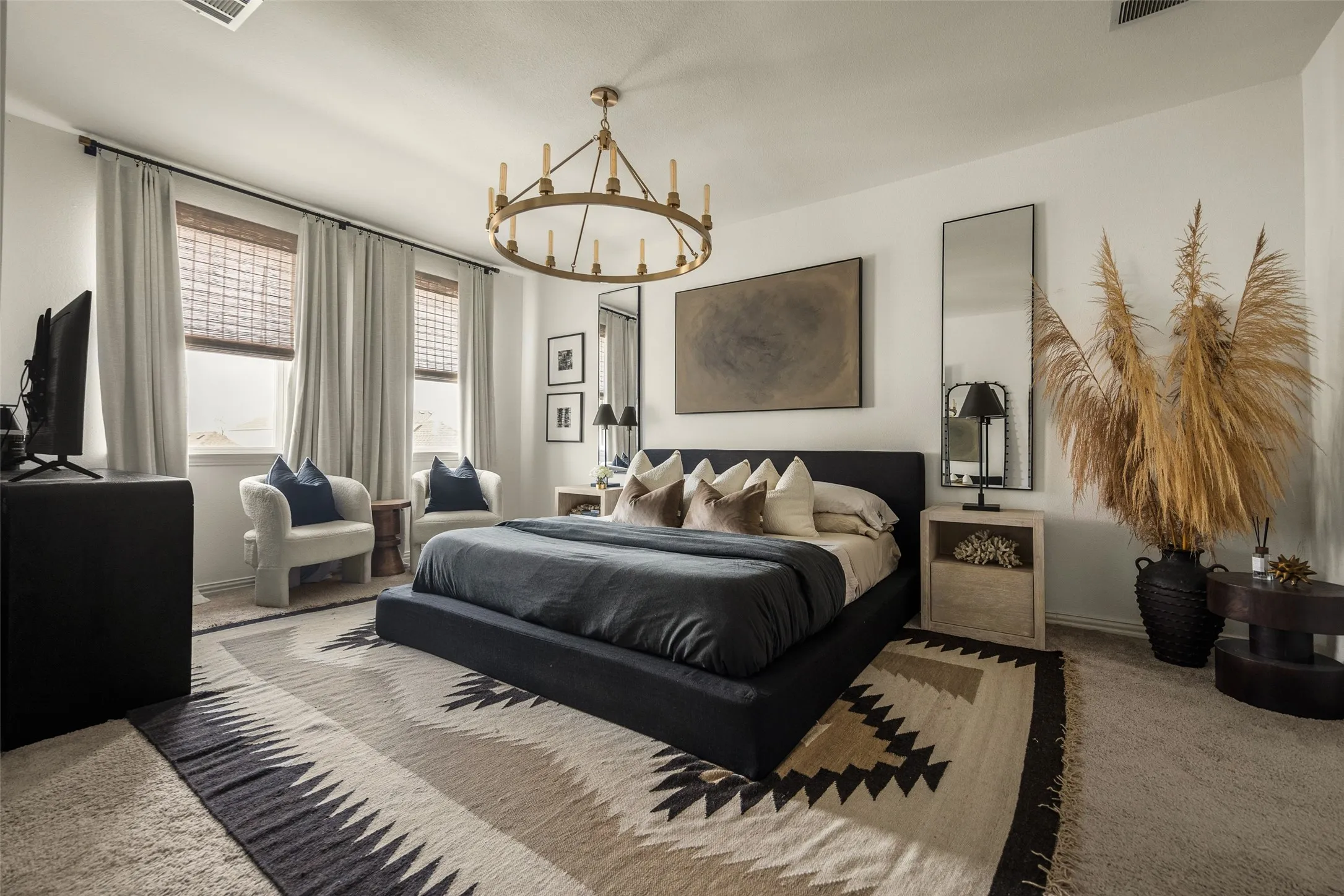 Recessed lighting, large windows with woven blinds and draperies, upholstered bed frame, and a prominent chandelier fixture