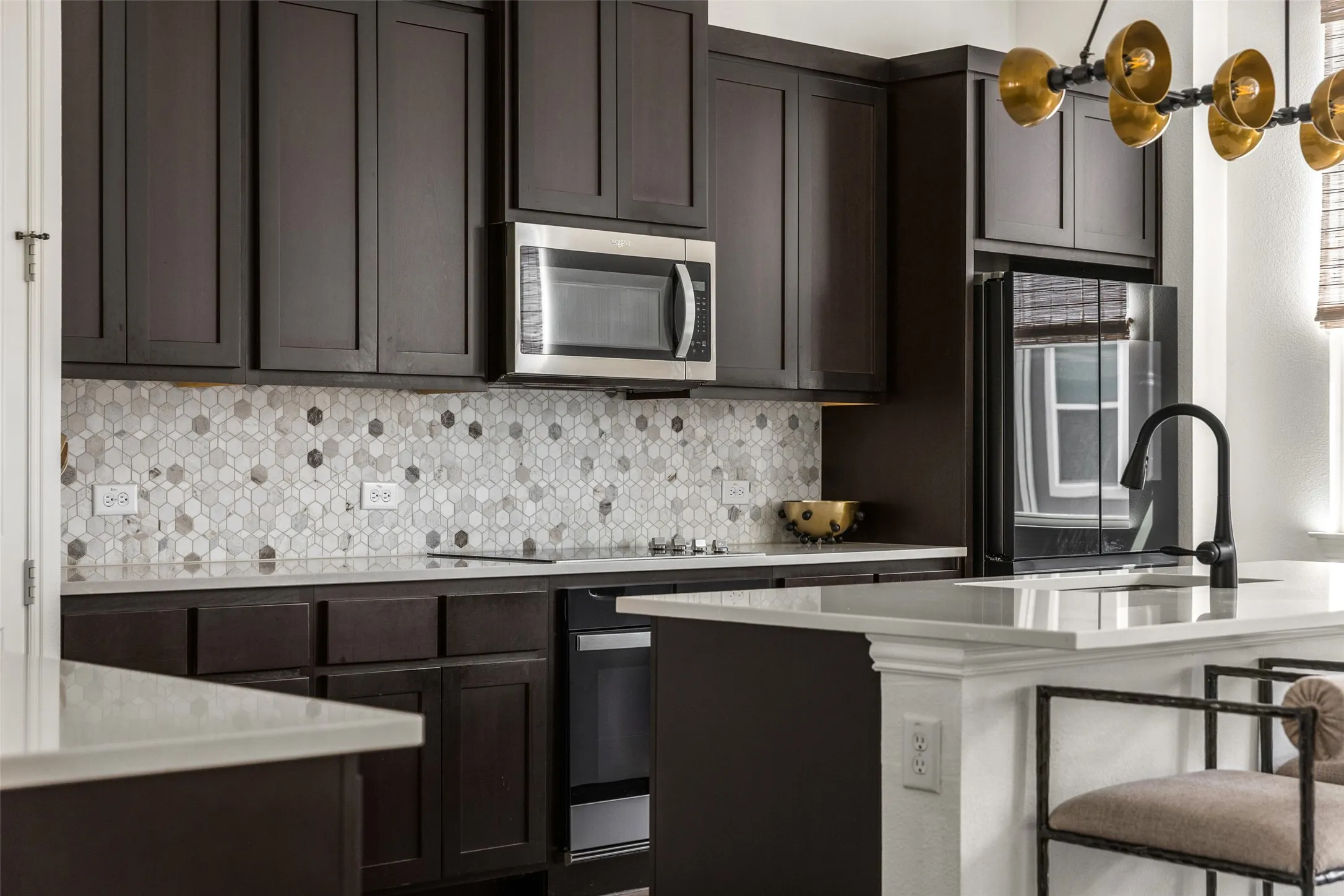 Dark wood-finish cabinetry, patterned tile backsplash, white countertops, built-in stainless steel microwave, and a black faucet