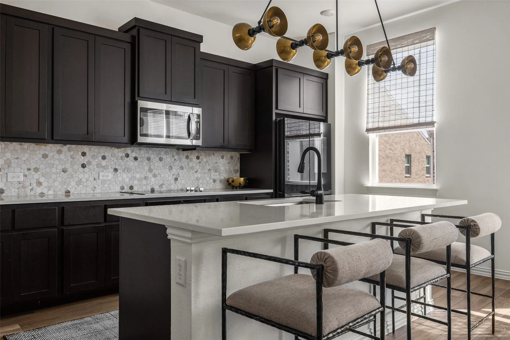 Dark wood-finish cabinetry, patterned tile backsplash, white countertop island, integrated sink with a black gooseneck faucet, and a large window with a woven shade