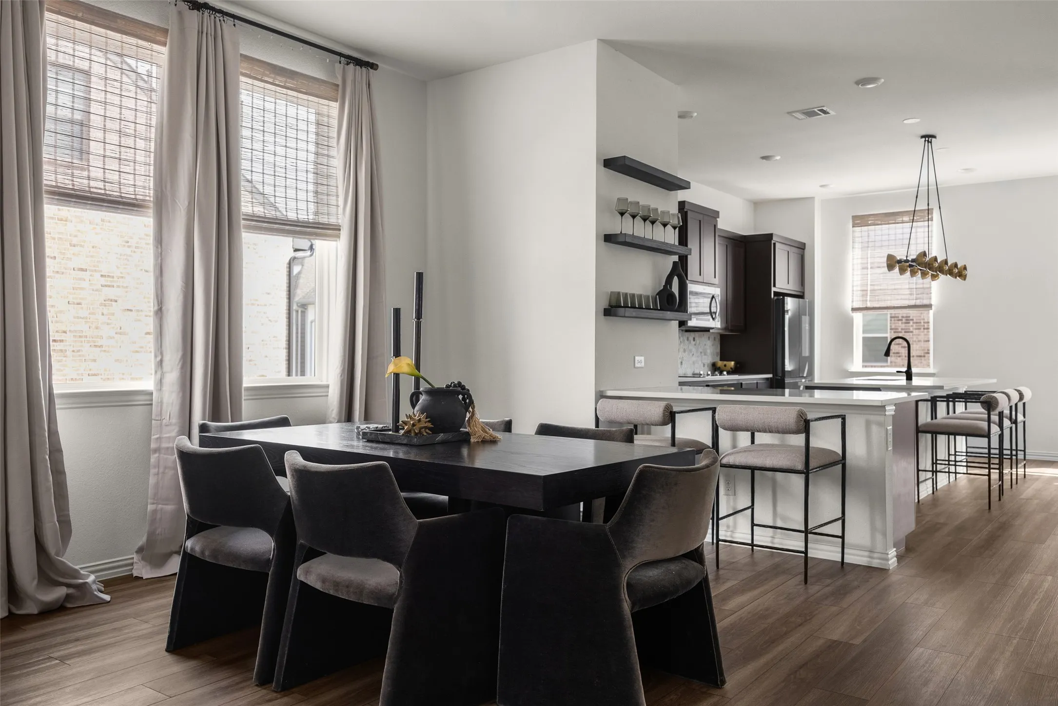 Open-concept living area featuring wood-finish flooring, a dining space with a built-in chandelier, kitchen island with seating, dark wood cabinetry, and open shelving
