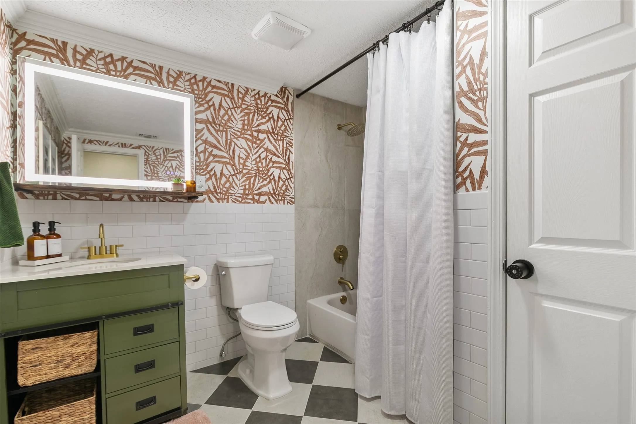 Bathroom featuring a green vanity with brass fixtures, an illuminated mirror, a toilet, a tiled shower, and patterned wallpaper