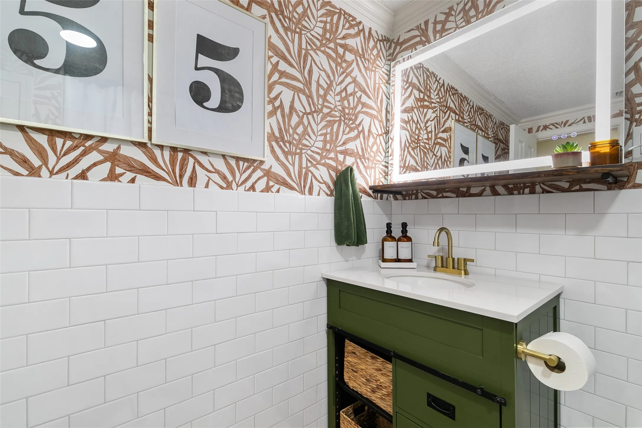 White subway tile wainscoting, green vanity with white countertop, gold-tone faucet, large framed mirror, and decorative wall covering