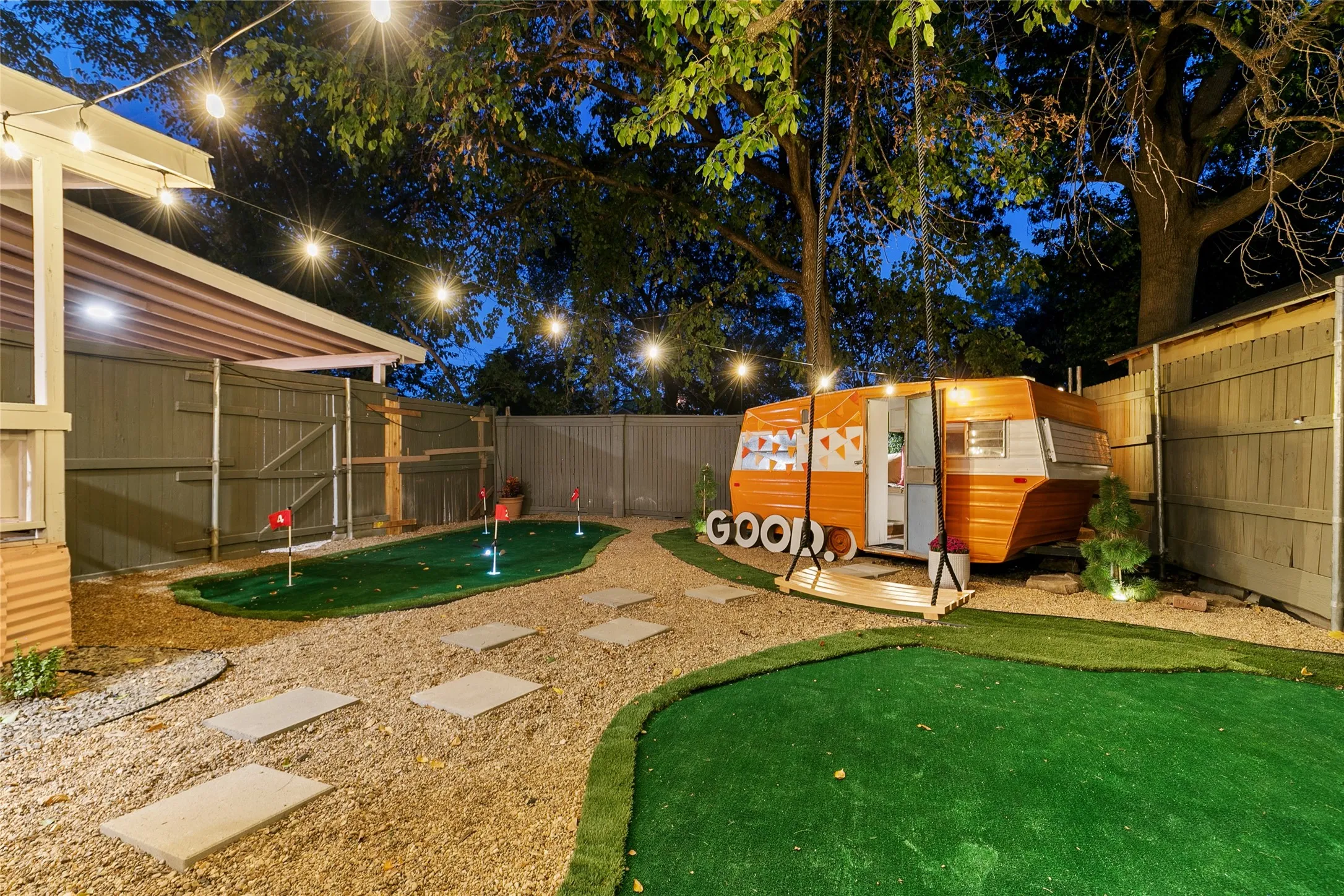 Putting green, gravel pathways with paver steps, string lighting, wood fencing, and a built-in swing