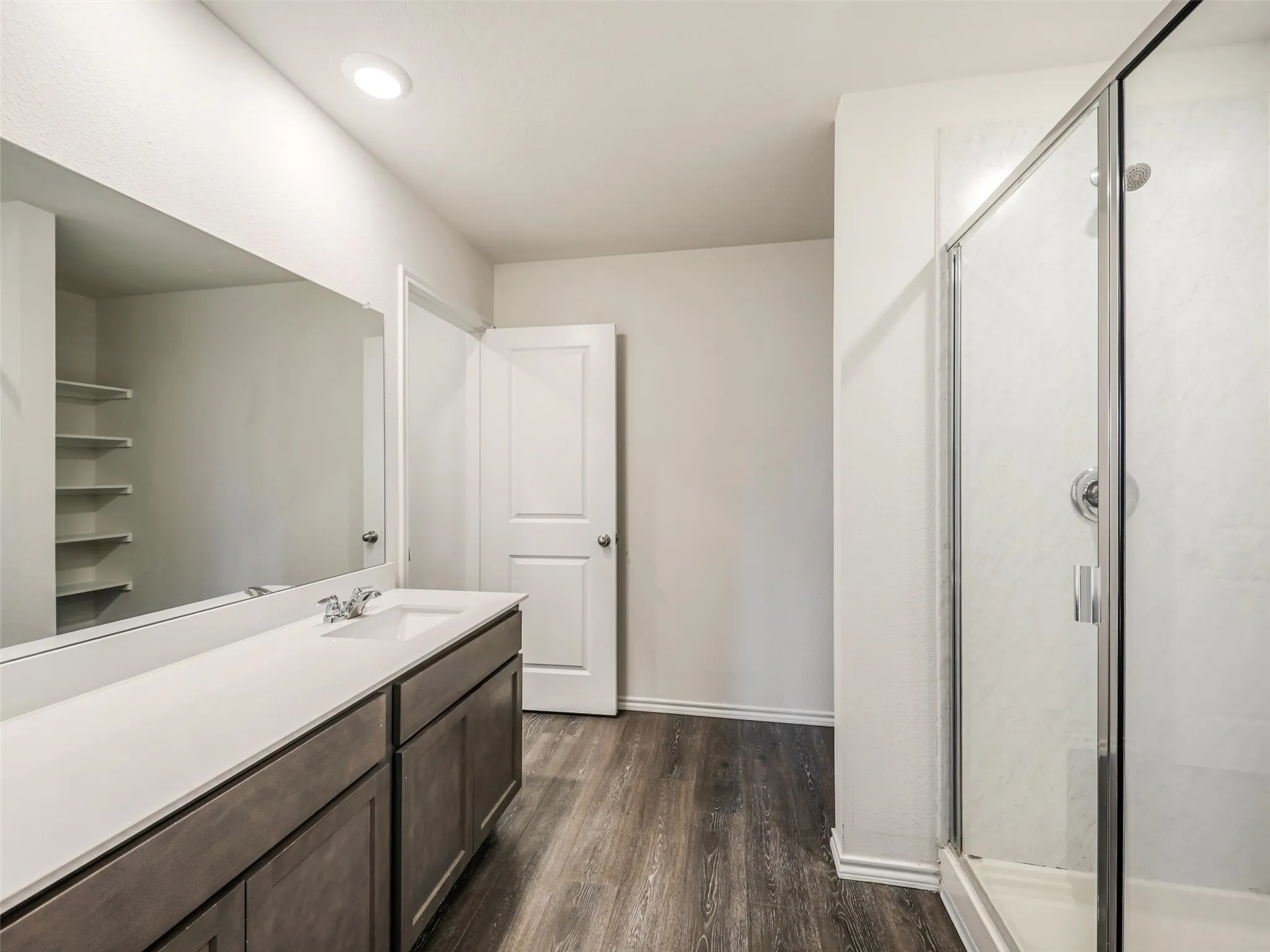 Spacious bathroom featuring a double vanity with a light countertop, wood-finish flooring, a frameless glass shower, and built-in shelving
