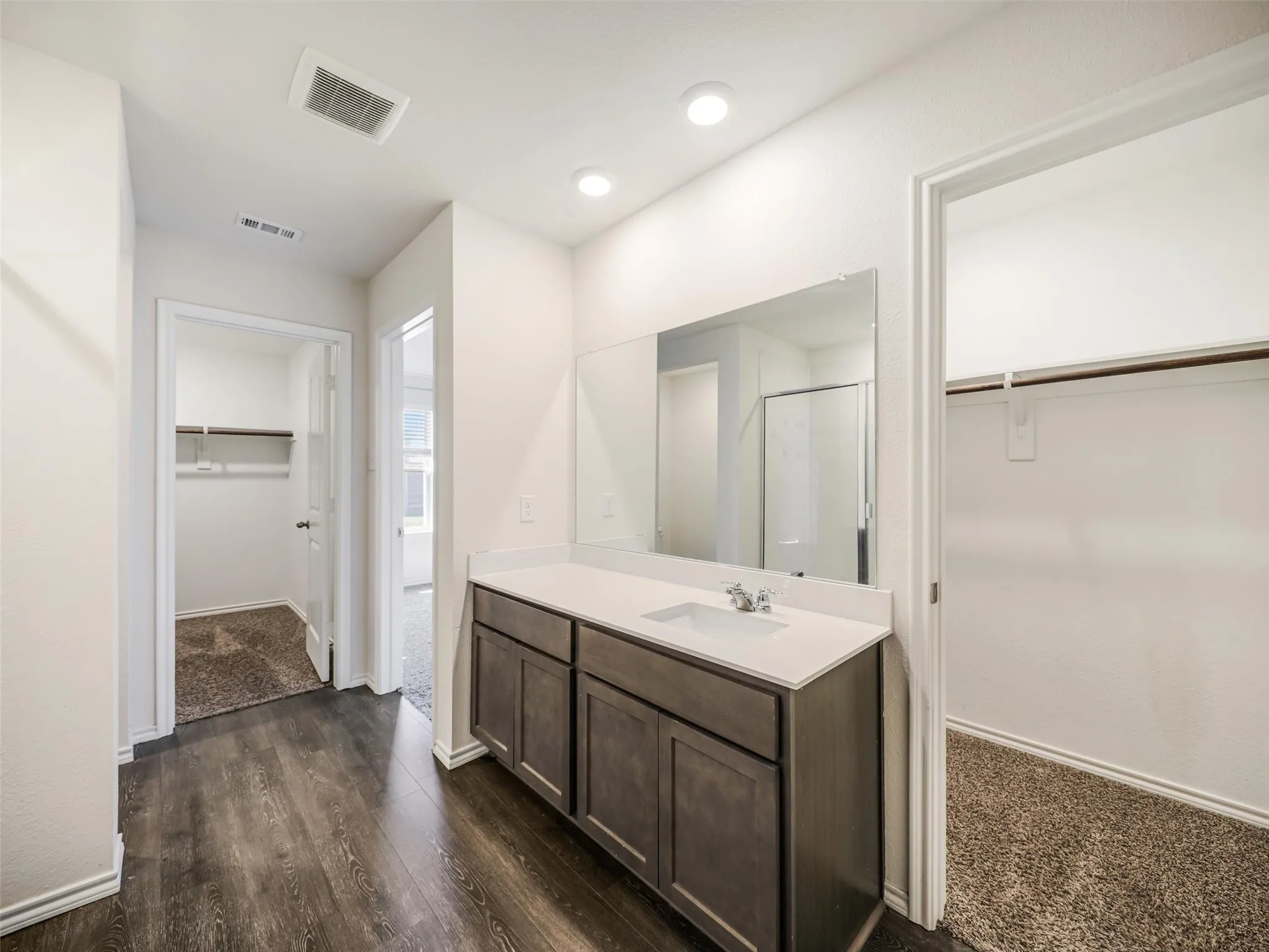 Spacious primary bathroom featuring a dark wood-finish vanity with dual sinks, white quartz countertop, large framed mirror, recessed ceiling lighting, and wood-finish flooring throughout