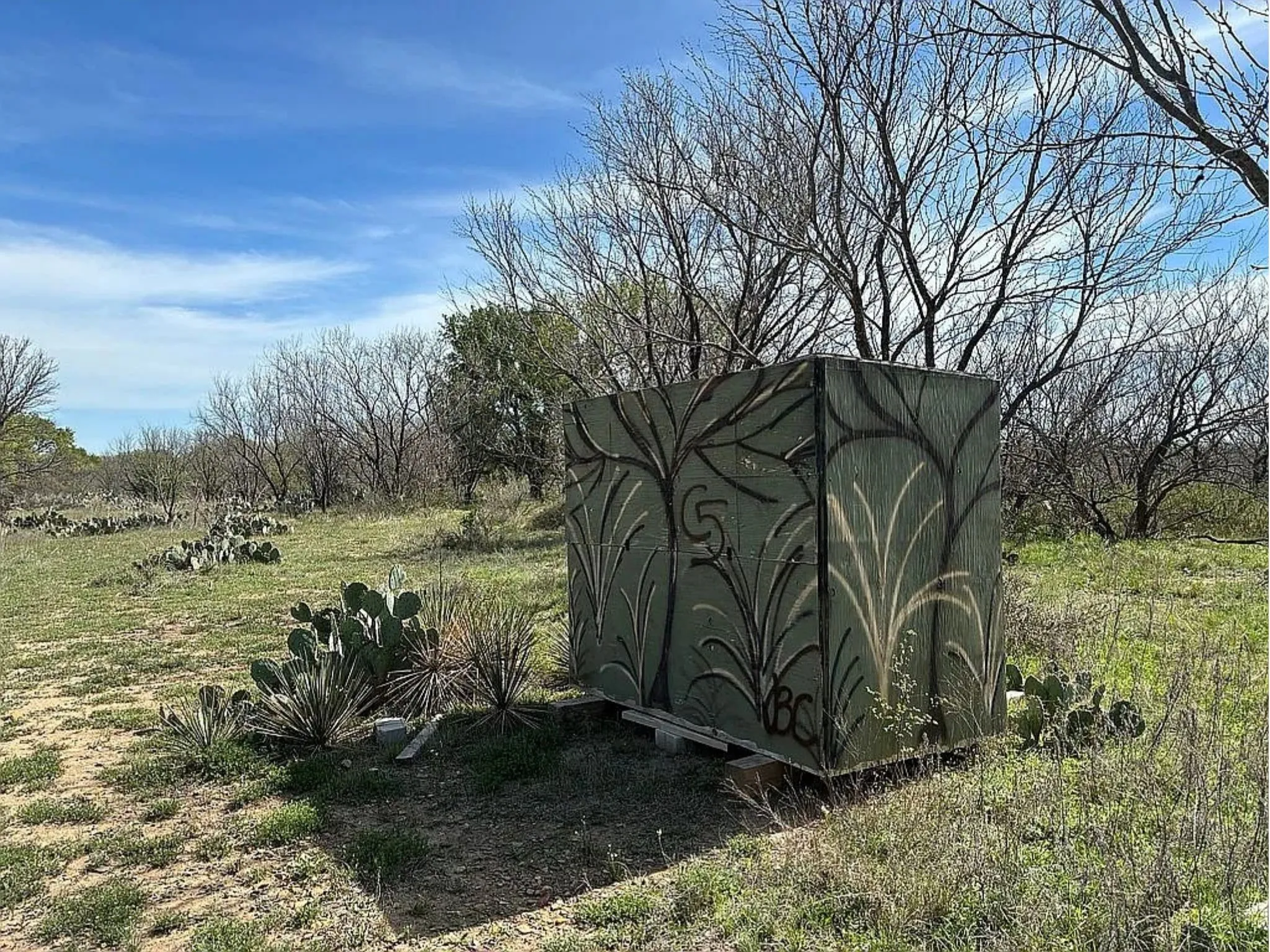 Additional contentThe image shows a decorative outdoor structure (appears to be a utility enclosure or garden feature) with ornamental relief panels in a rural or semi-rural setting with sparse vegetation, bare trees, and open grassland