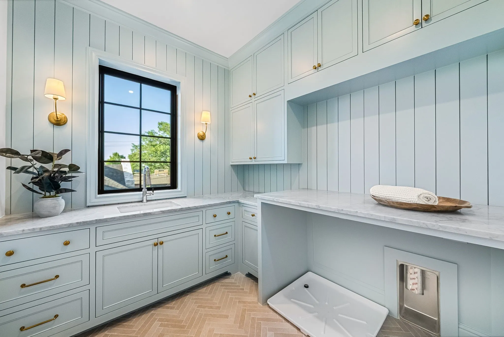 Built-in cabinetry with brass hardware, marble countertops, window over undermount sink with gooseneck faucet, full-height paneled walls, and herringbone wood-finish flooring