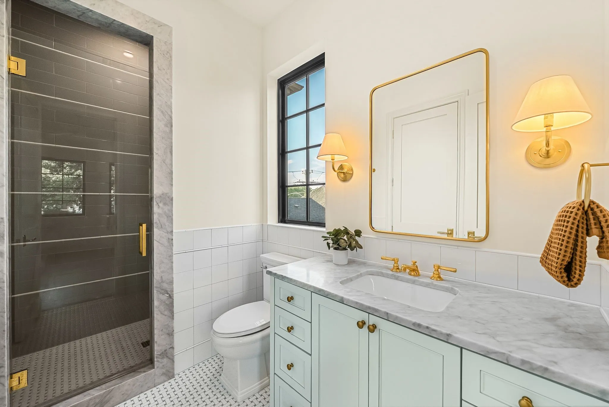 Glass-enclosed shower with marble-look surround, light blue vanity with gold hardware, white rectangular sink, gold-framed mirror, and patterned tile flooring