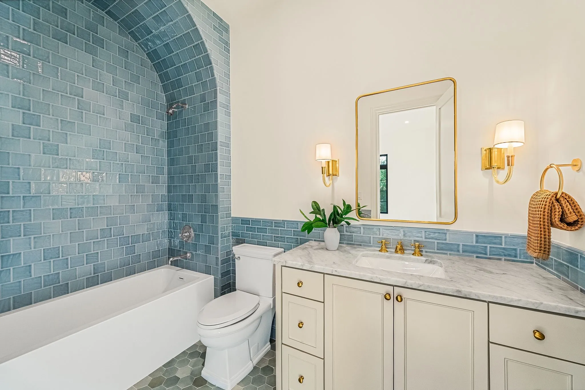 White bathtub with an arched blue subway tile surround, hexagonal floor tiling, a white toilet, and a vanity with a white countertop and brass hardware
