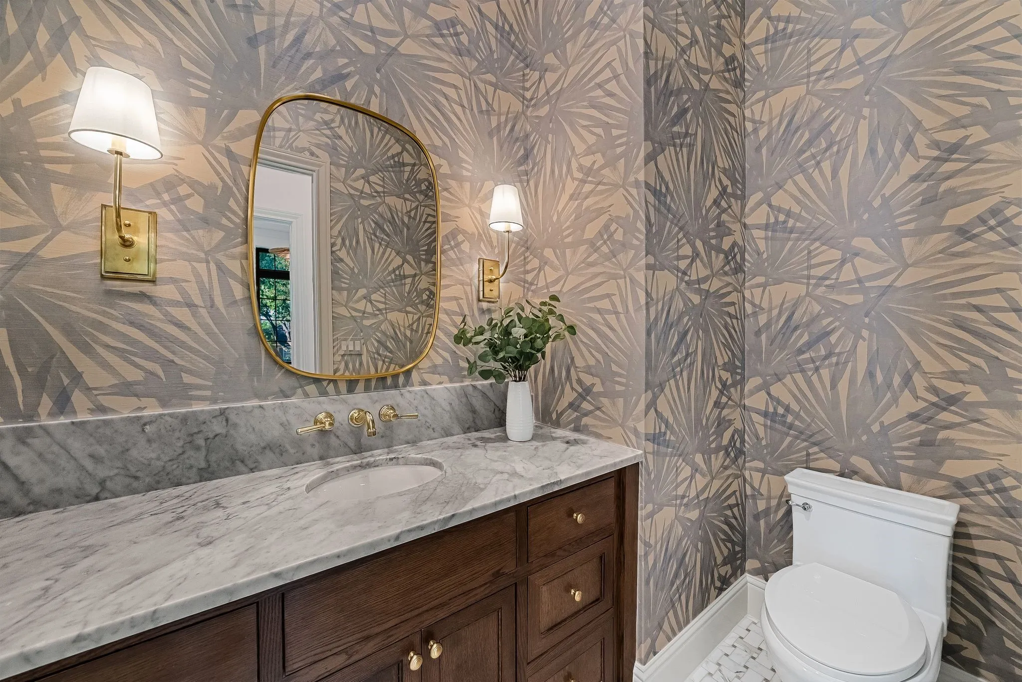 Spa-inspired powder room featuring a marble-topped double vanity with brass hardware and faucet, arched brass-framed mirror, patterned wallcovering with botanical motifs, brass wall sconces, and a white porcelain toilet with matching tile accents