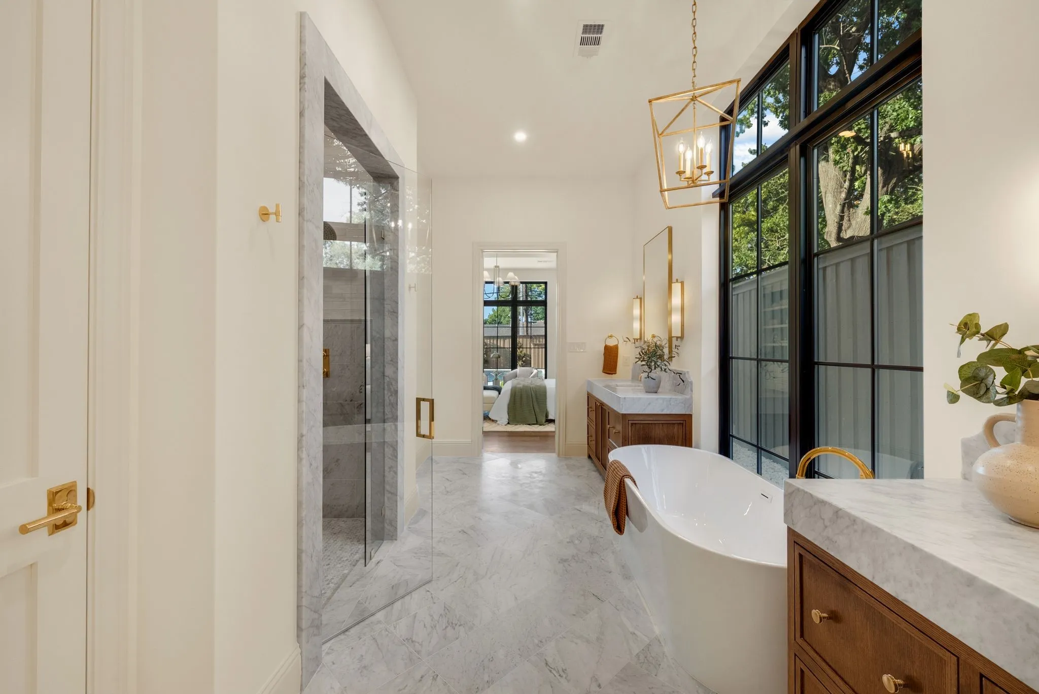 Spacious bathroom featuring marble-finish flooring, a freestanding soaking tub, a large window with black framing, a double vanity with wood-finish cabinetry, and a walk-in shower with glass enclosure