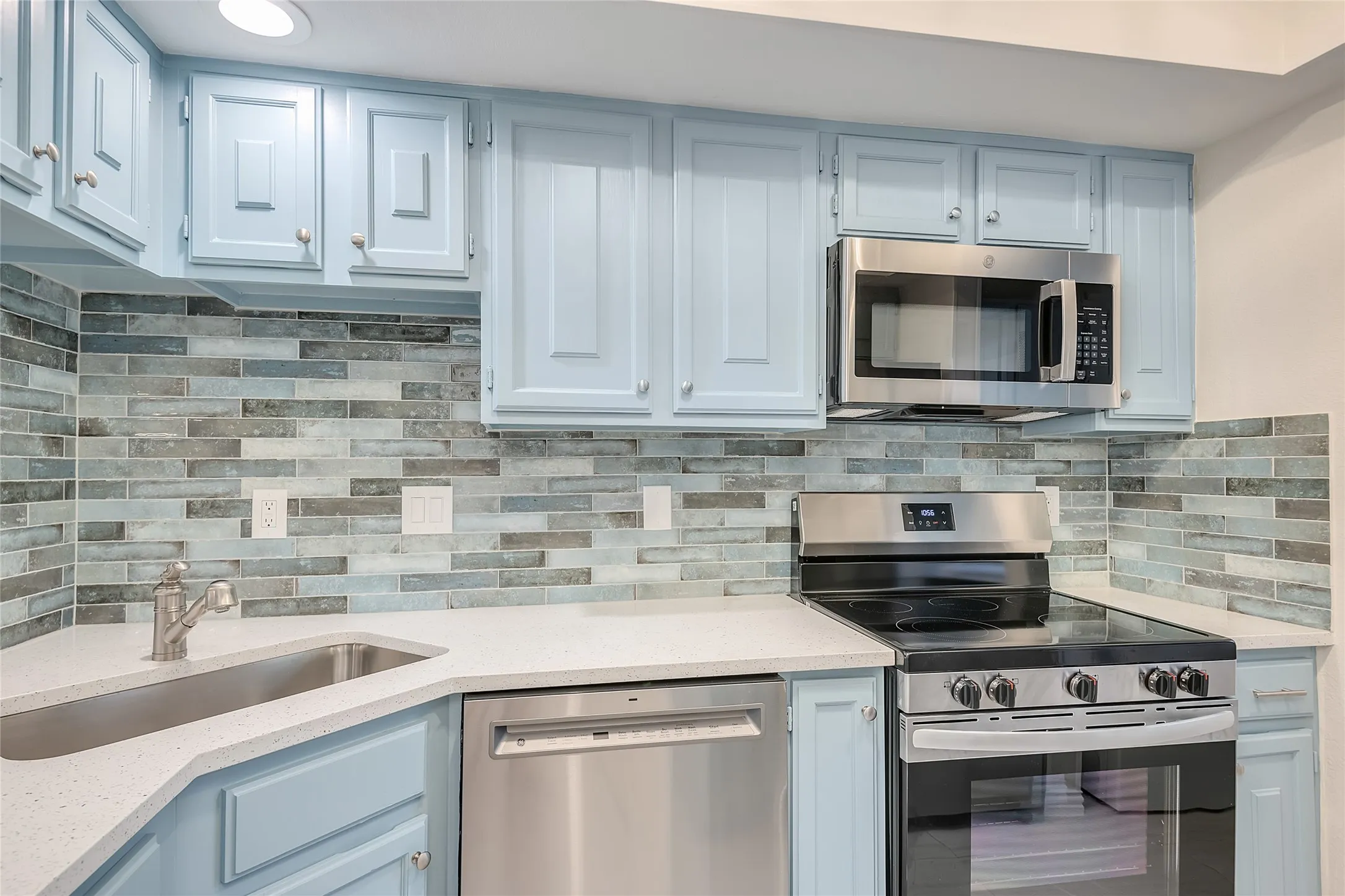 Kitchen featuring light blue cabinetry, stainless steel appliances, a tile backsplash, light-toned countertops, and a corner sink