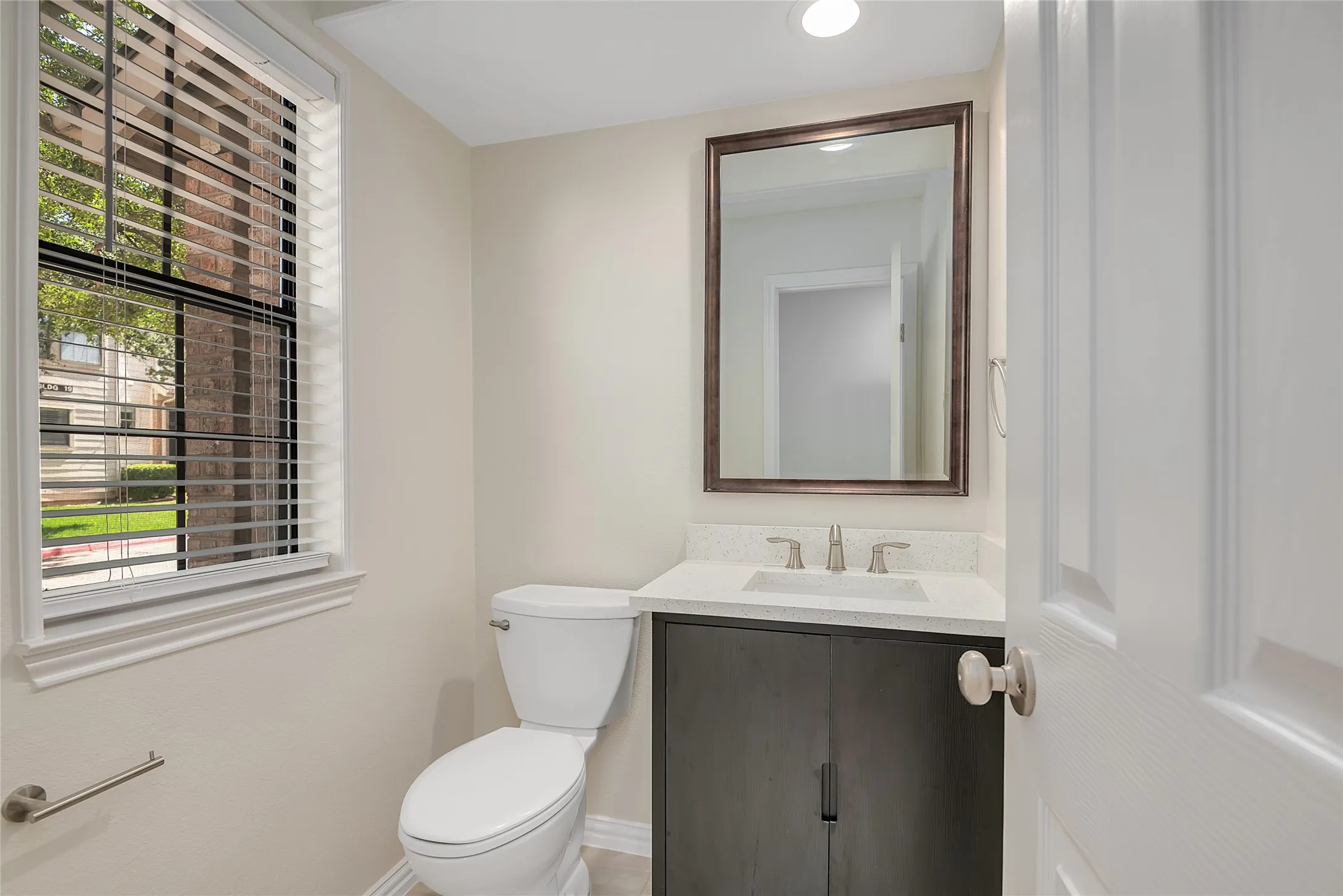 Recessed lighting, framed mirror, wood-finish vanity with a light-toned countertop, and a window with horizontal blinds