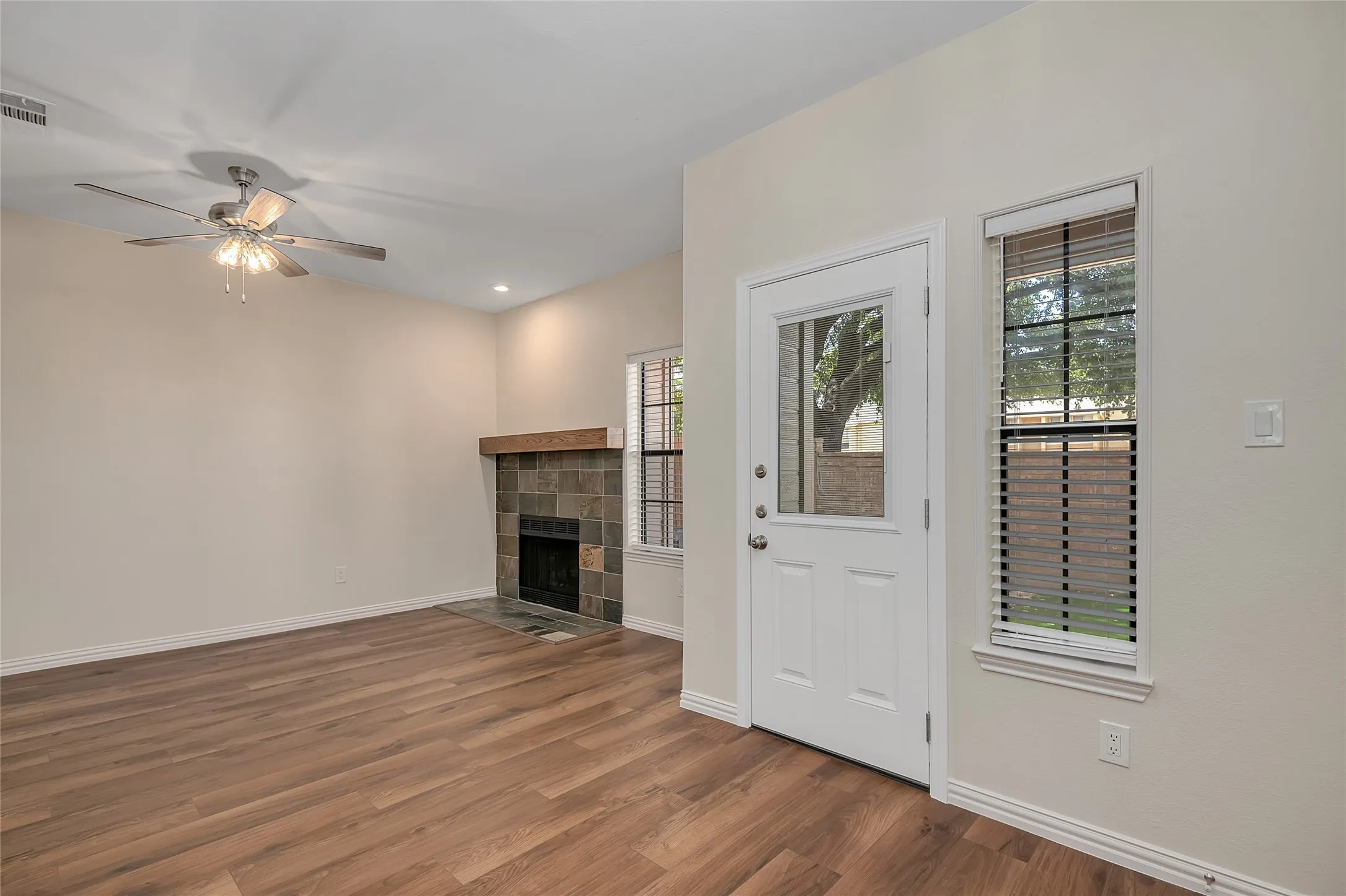 Wood-finish flooring, tiled fireplace with a wood mantle, ceiling fan, white paneled door with glass insert, and a window with blinds