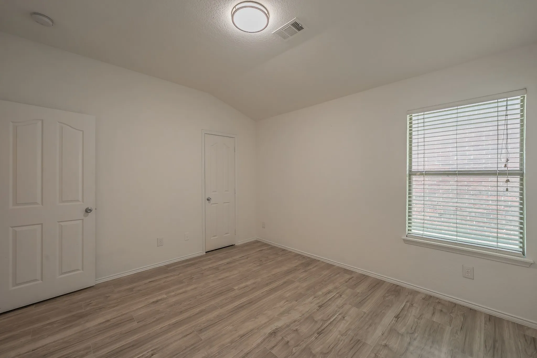 Vaulted ceiling, wood-finish flooring, white walls, window with blinds, and flush-mount ceiling light