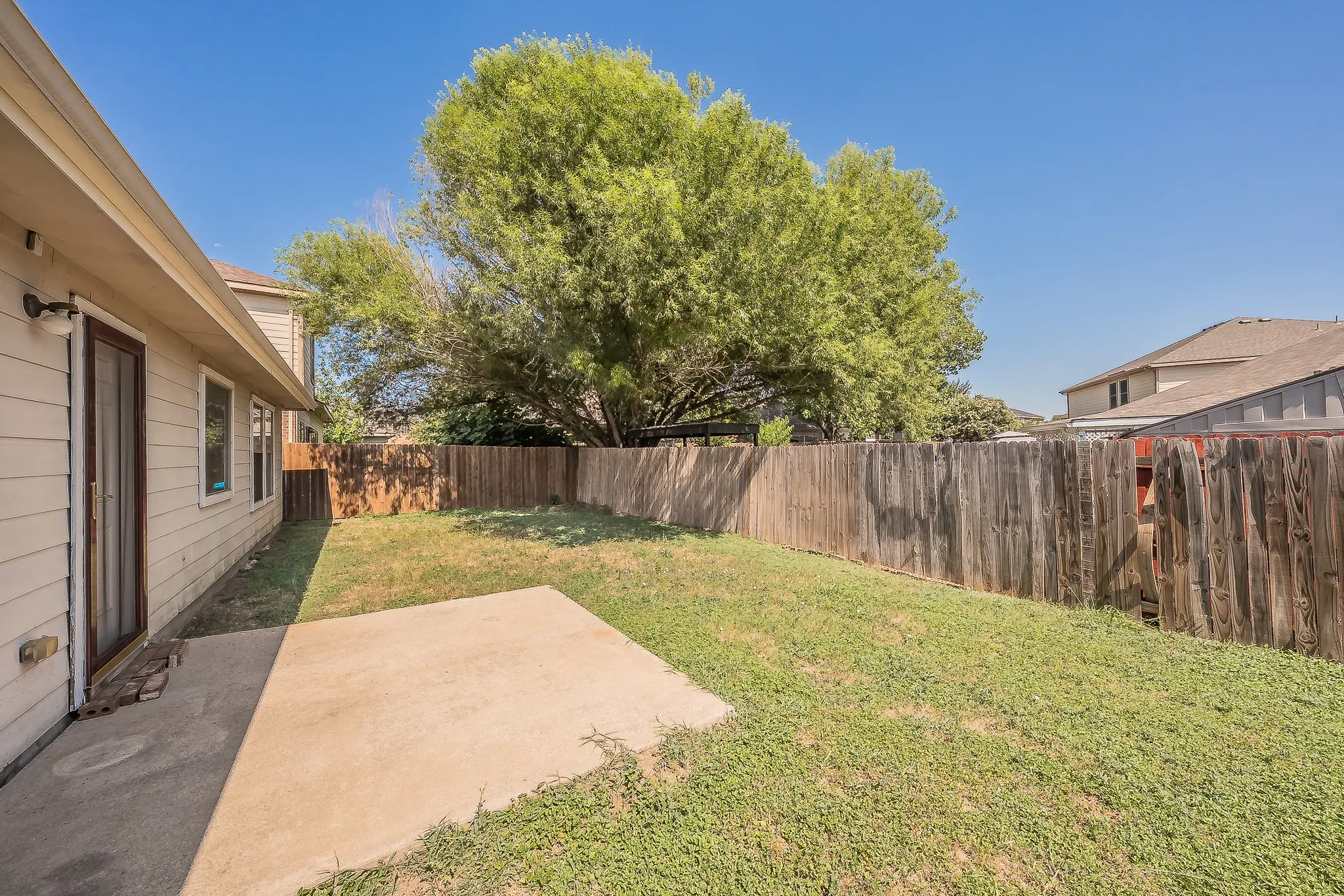 Concrete patio with a sliding glass door, leading to a fenced backyard with a mature tree
