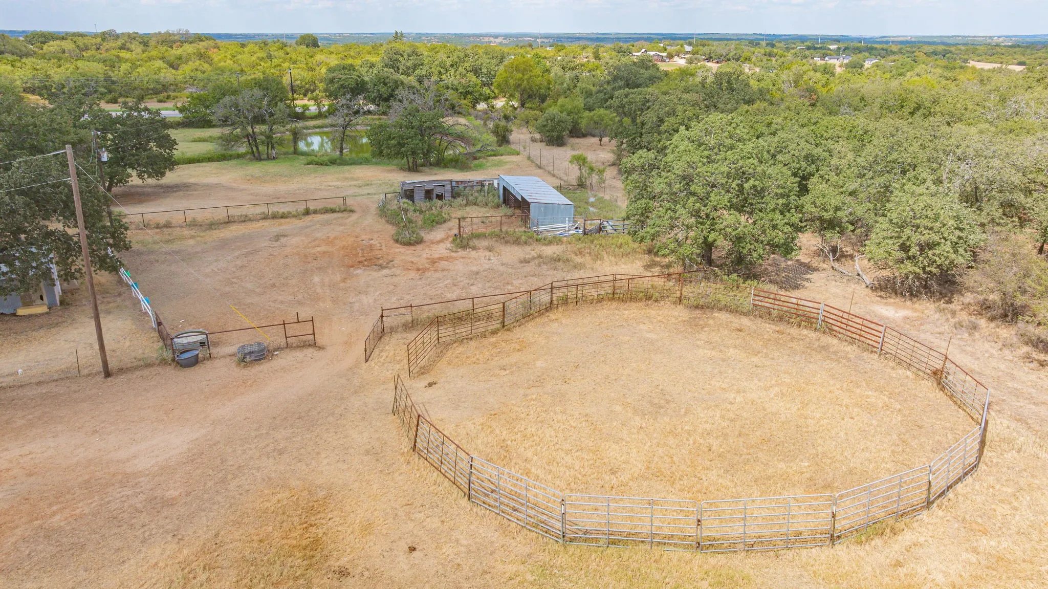 Circular fenced arena, multiple fenced areas, outbuilding with corrugated metal roof, mature trees, and a visible pond