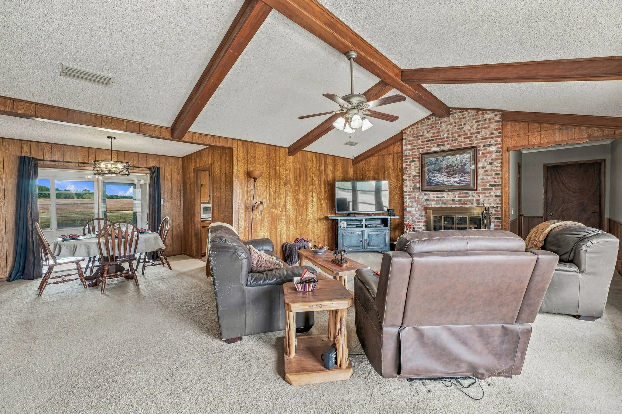 Vaulted ceiling with wood beams, brick fireplace, wood-panel walls, ceiling fan, and carpet flooring