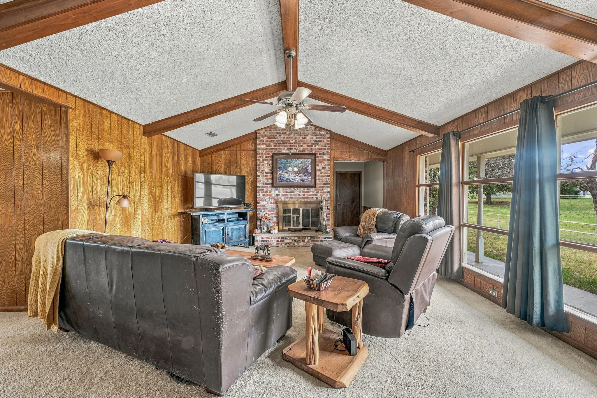 Vaulted ceiling with exposed wood beams, brick fireplace, wood paneling, and large windows