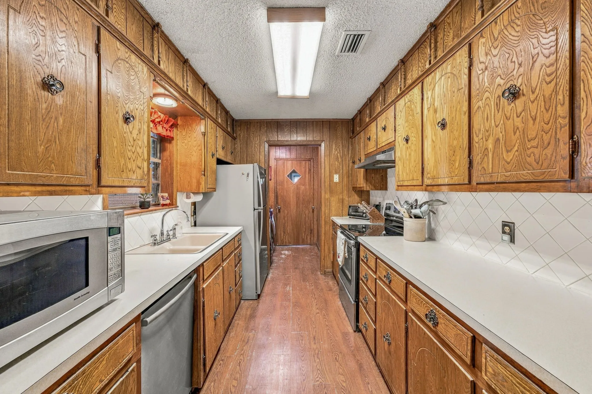 Galley kitchen featuring rich wood cabinetry with detailed paneling, stainless steel appliances including a refrigerator and electric range, white subway tile backsplash, dual undermount sinks with chrome fixtures, and wood-finish flooring throughout