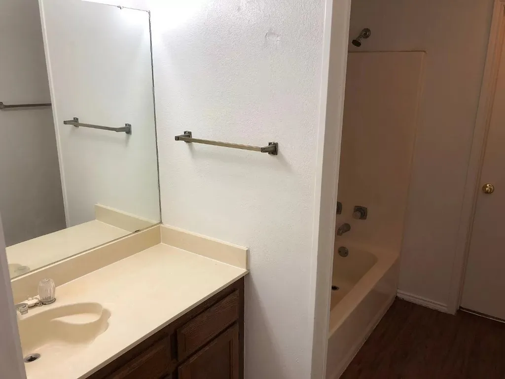 Vanity with integrated sink, full-width mirror, wood-finish cabinetry, and a full bathtub with shower fixture
