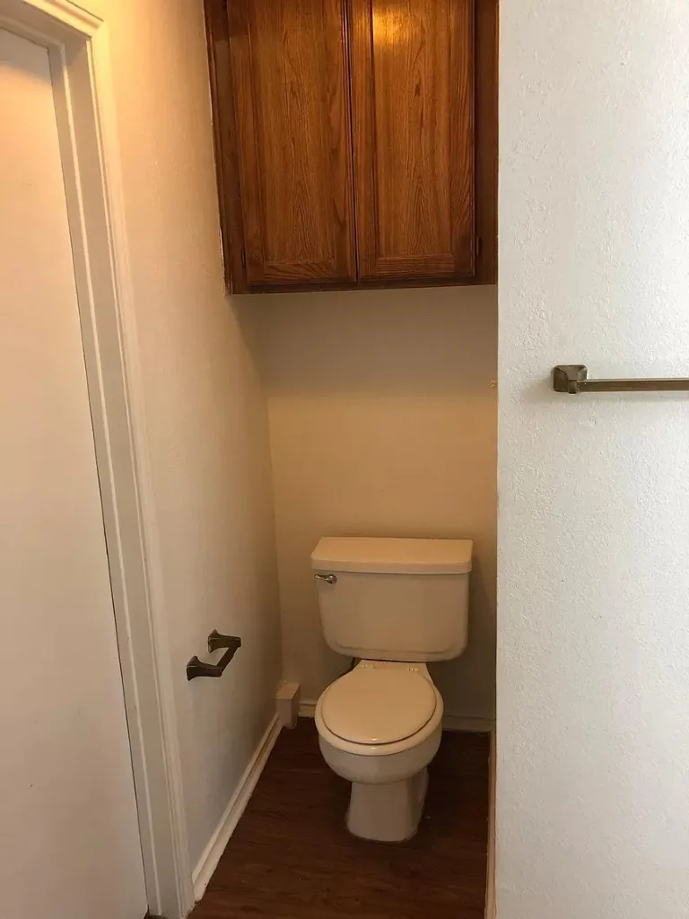 White toilet, wood-finish flooring, and overhead cabinetry