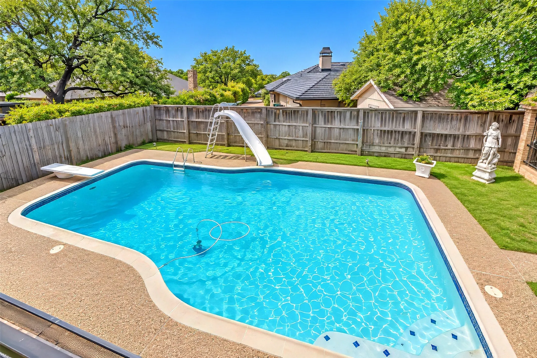 Rectangular swimming pool featuring a diving board and attached slide, surrounded by a textured concrete deck and a wooden privacy fence