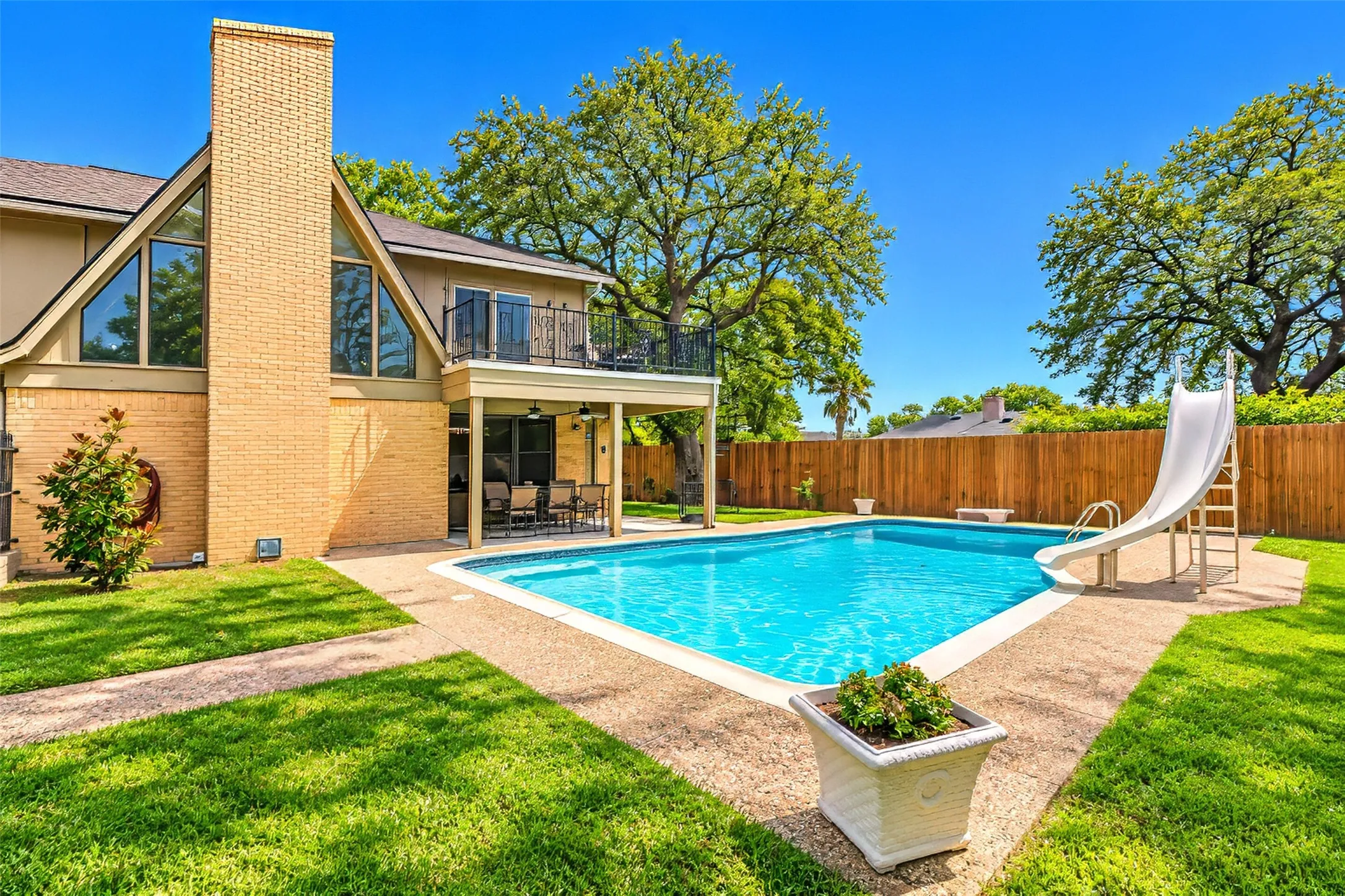 In-ground swimming pool, brick chimney, gabled windows, upper-level balcony with iron railing, covered patio