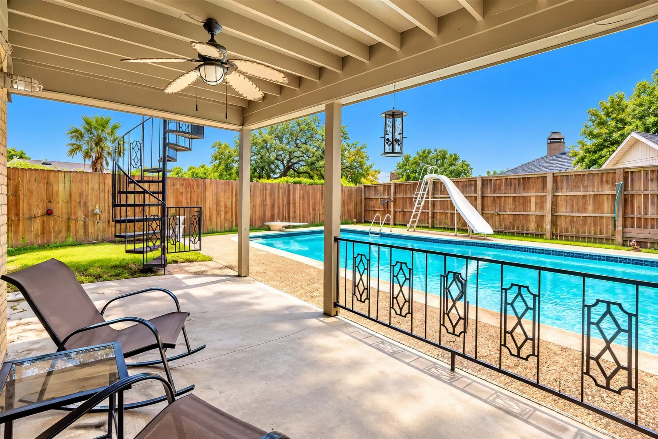 Covered patio with ceiling fan, spiral staircase, and full-length swimming pool with slide, enclosed by a wood privacy fence