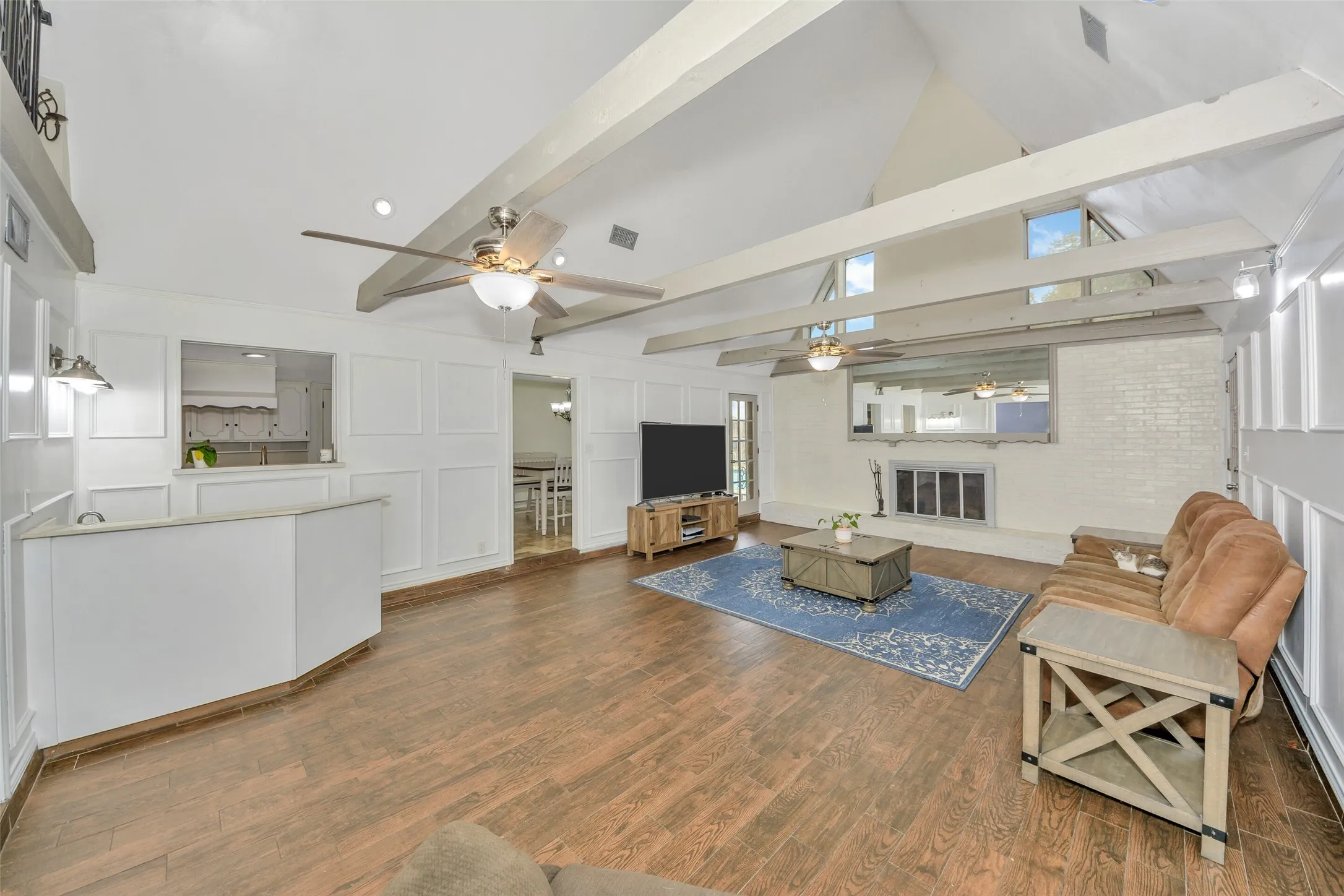 Vaulted ceiling with exposed beams, wood-finish flooring, white paneled wall accents, white brick fireplace, and pass-through counter