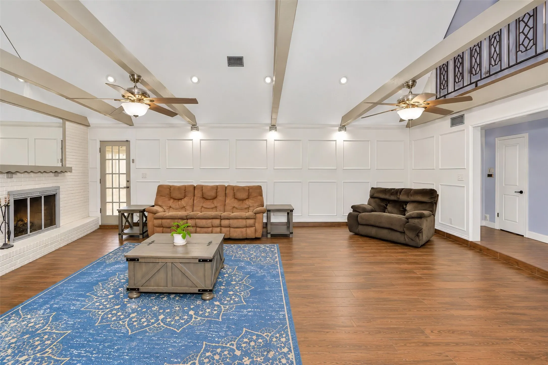 Vaulted ceilings with exposed beams, wood-finish flooring, white brick fireplace, decorative wainscoting, and recessed lighting