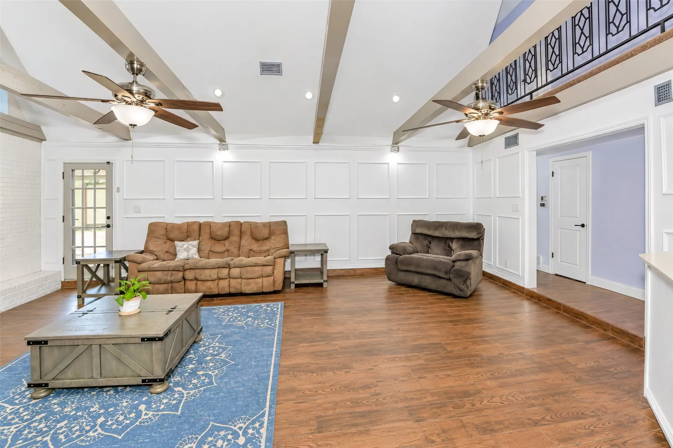 High vaulted ceiling with exposed beams, wood-finish flooring, white paneled wall accents, recessed lighting, and a second-story interior balcony with an ornate railing
